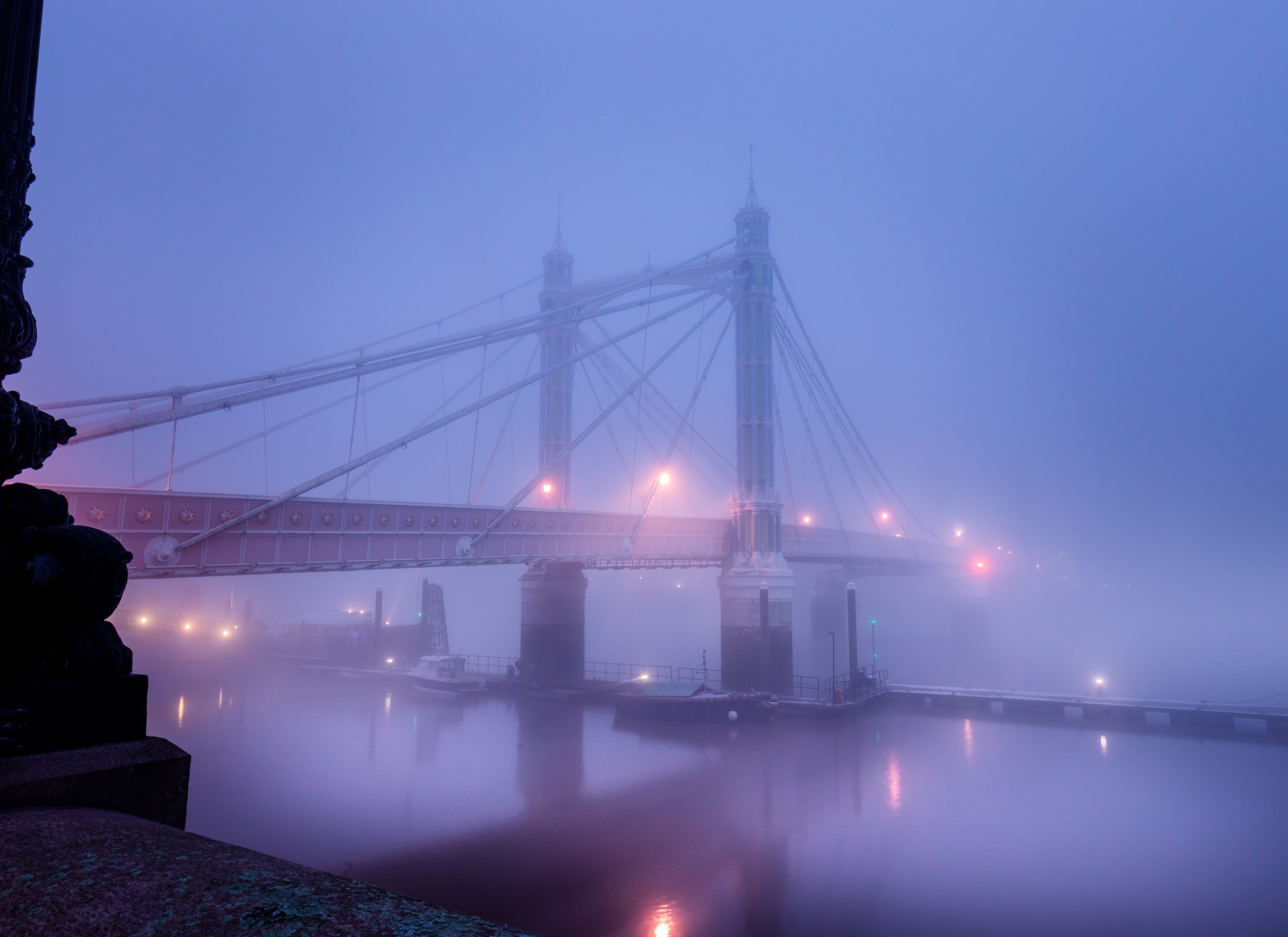 Suspension bridge shrouded in fog with dimly lit lights, reflecting in calm water.