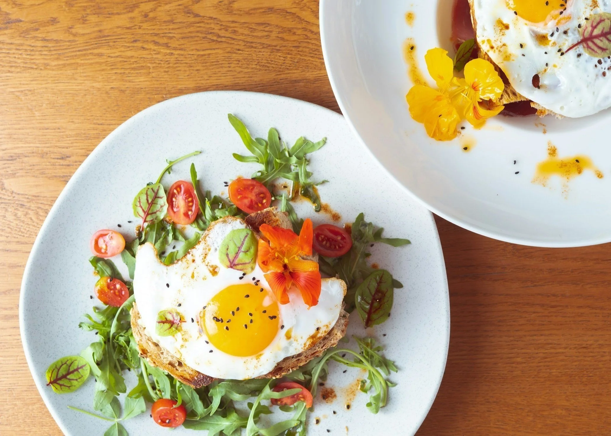 Eggs on toasted bread with arugula, cherry tomatoes, and edible flowers on white plates.