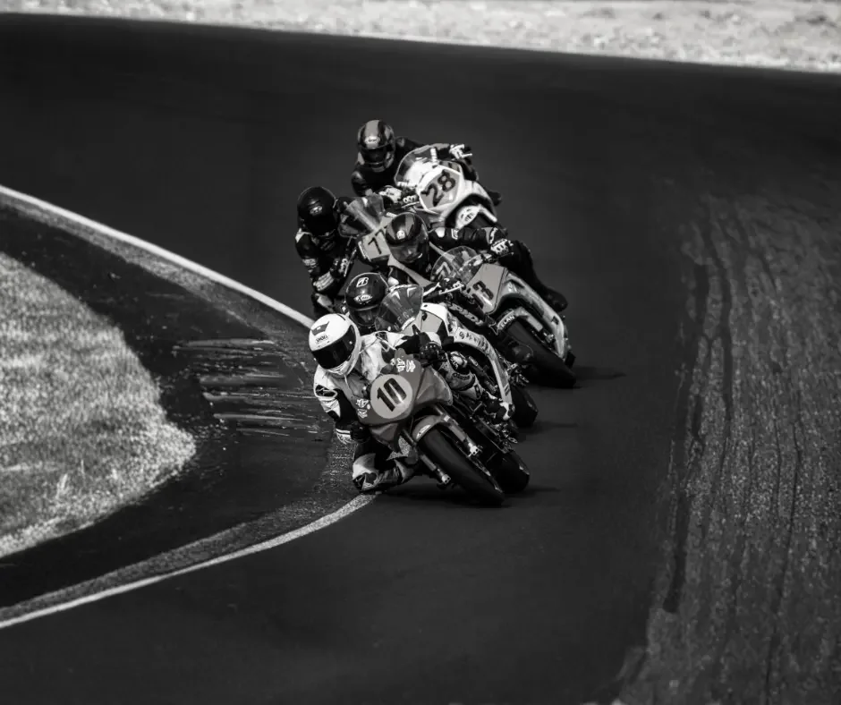 A man in an orange safety jacket is kneeling on grass, preparing a large drone on a wooden platform. There is a black case nearby, and a background of blue sky with clouds and distant trees and buildings.