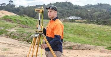 Surveyor standing on construction site with tripod and surveying equipment, outdoors on cloudy day.
