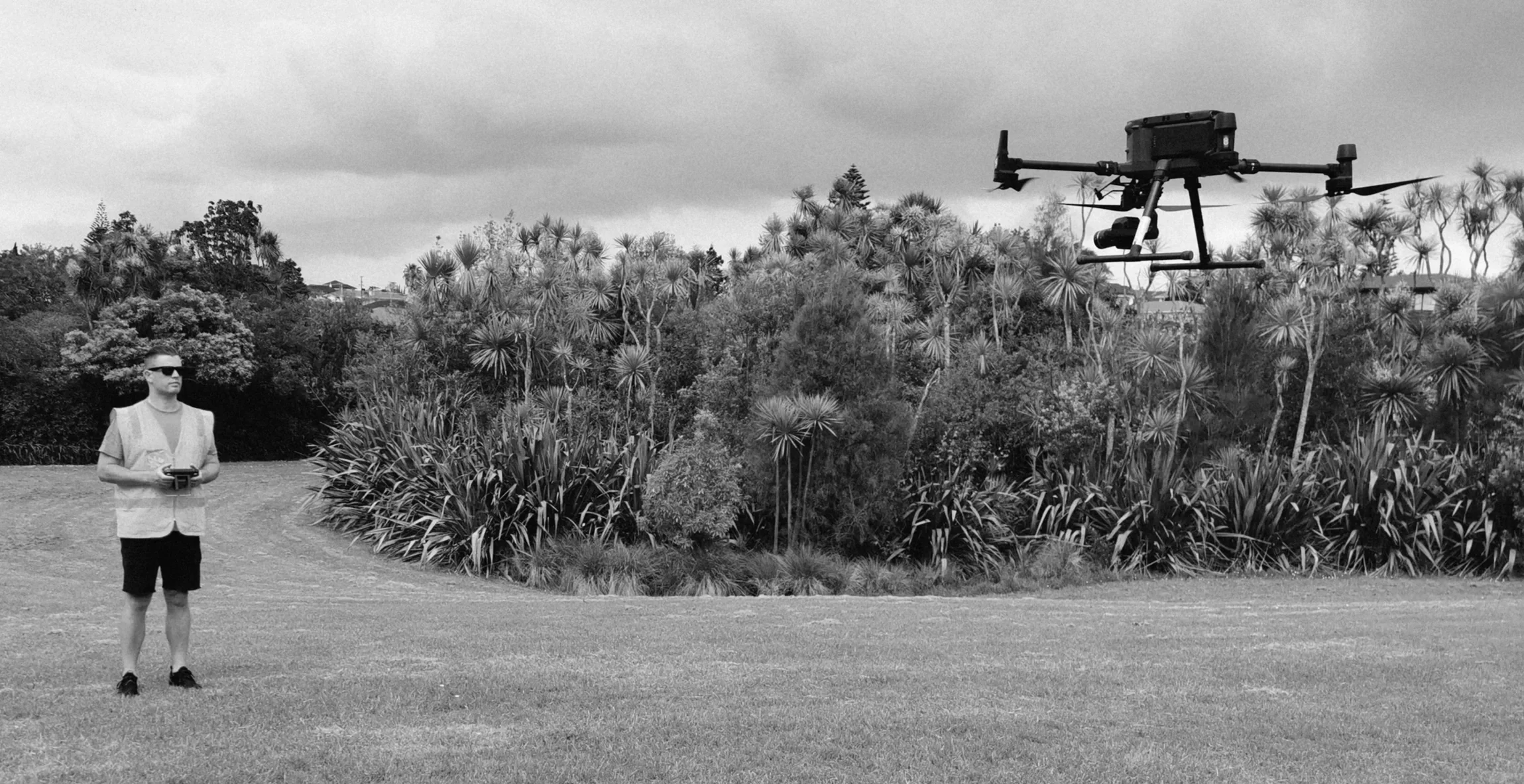 A man in sunglasses and an orange safety vest is standing on a grassy field, controlling a drone with a remote. The drone is flying in the air against a cloudy sky, surrounded by trees and bushes.