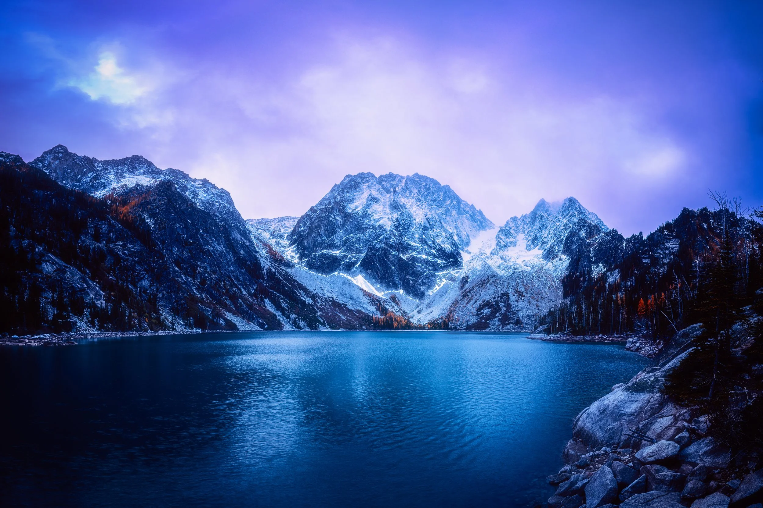 Snow-capped mountains surrounding a calm alpine lake under a cloudy sky. By Archie Carmel.