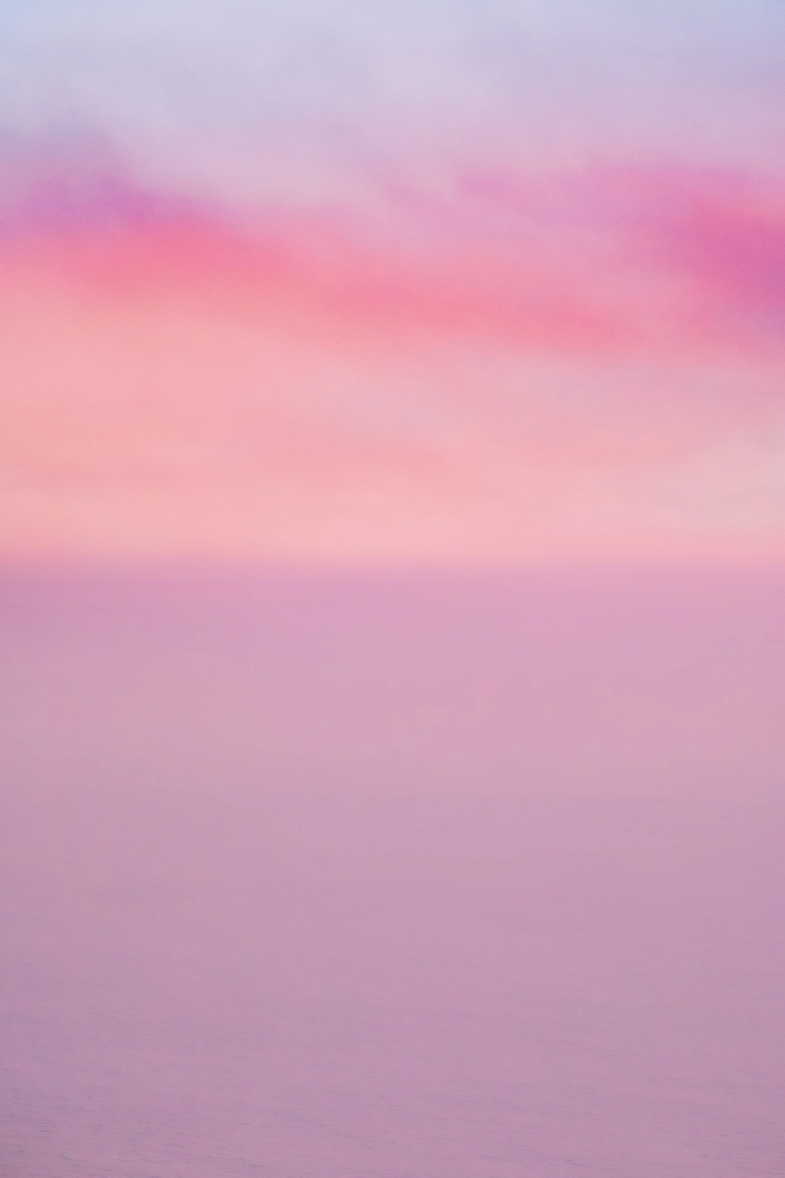 Sunset over a calm pink ocean with soft waves. By Archie Carmel.