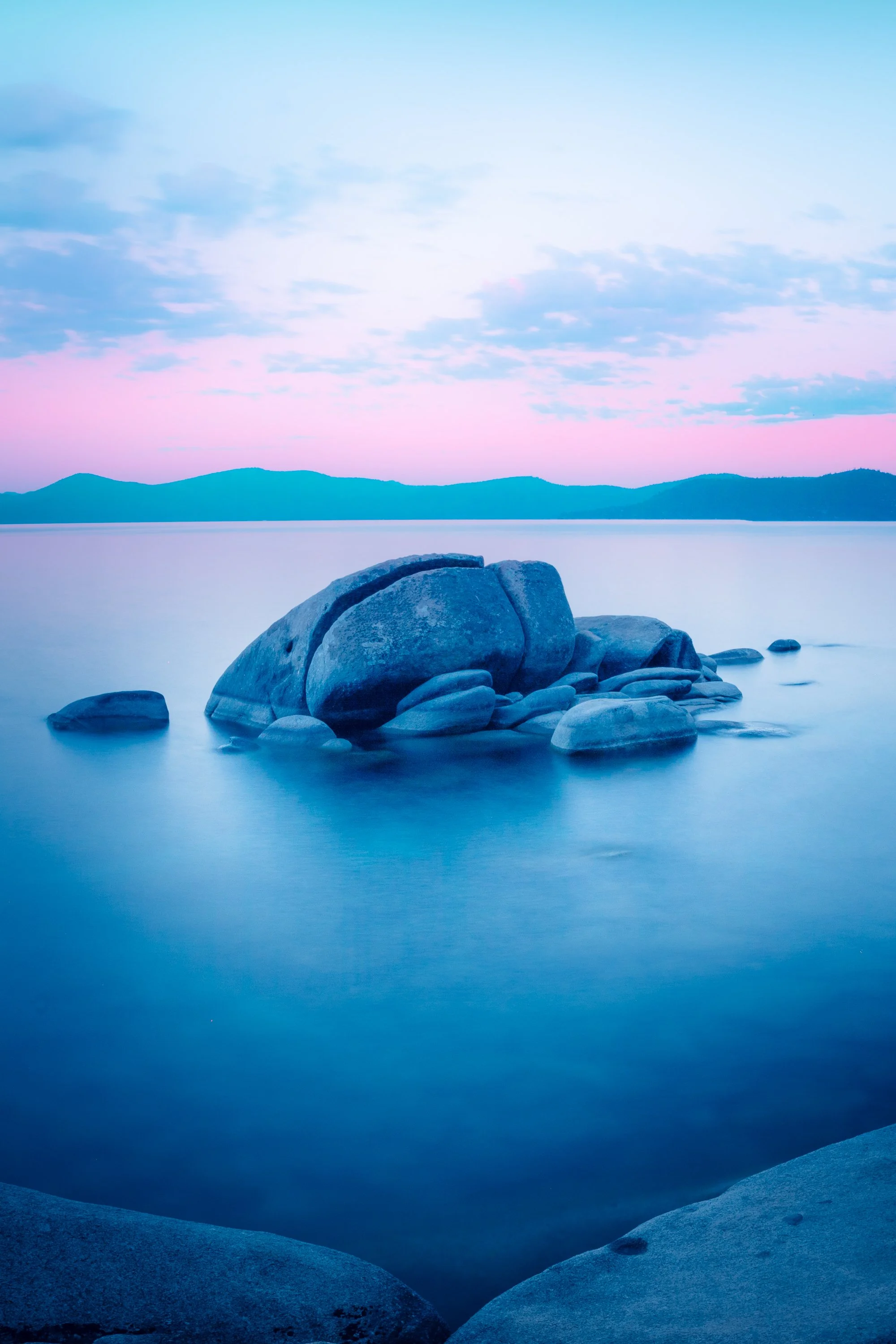 A soft pink sunrise over Lake Tahoe with reflections of soft blues and pinks over the glassy lake. By Archie Carmel.