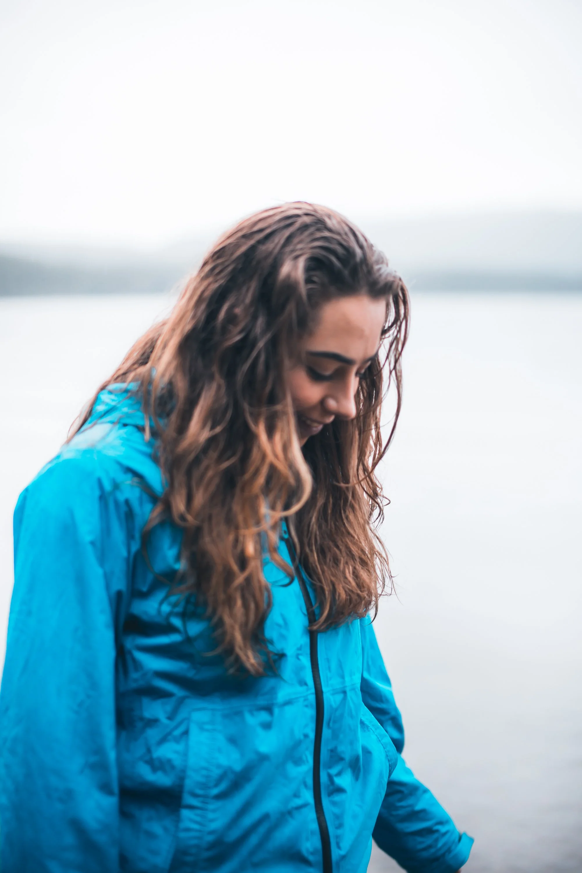 A woman with long dark hair and blue eyes, wearing a black top and a brown strap across her shoulder, standing outdoors with a blurred natural background. Portrait by Archie Carmel.