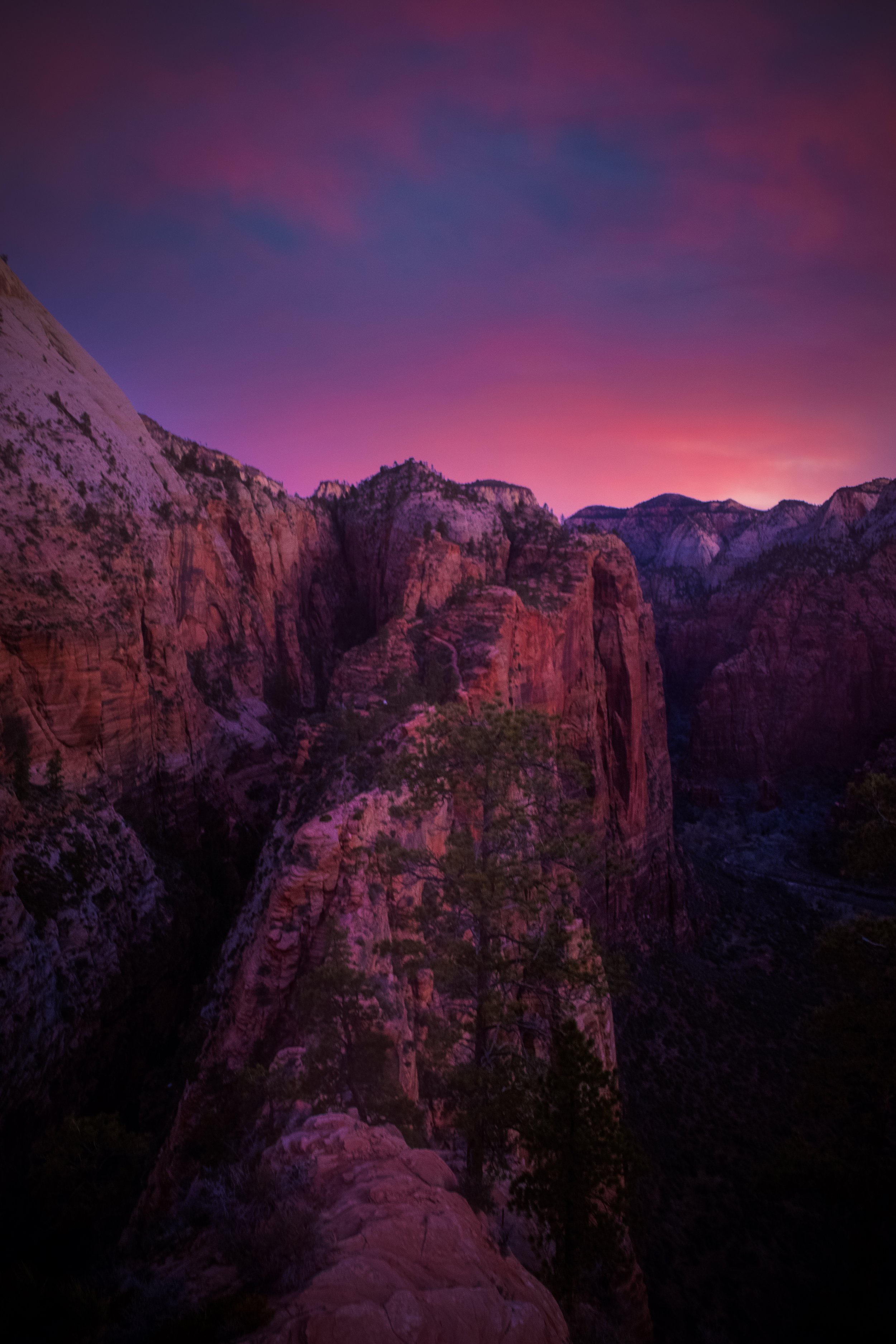 A mountain landscape during sunset with colorful pink and purple sky, rugged rock formations, and sparse trees. By Archie Carmel.