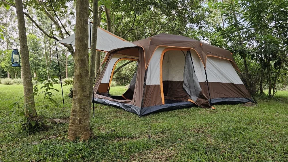 A large camping tent set up in a grassy area surrounded by trees, with an attached canopy and a small lantern hanging from a nearby tree.