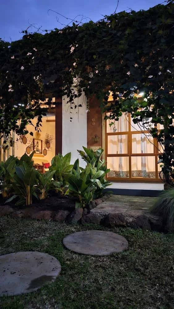 A house with large windows glowing with warm light, surrounded by lush green plants and a canopy of leaves overhead, with stepping stones on the lawn outside.