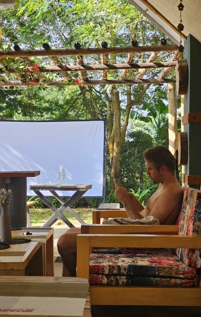 A man sitting on a cushioned wooden chair with a floral pattern, looking at his phone, in an outdoor patio area with lush greenery and trees, sunlight filtering through the leaves.