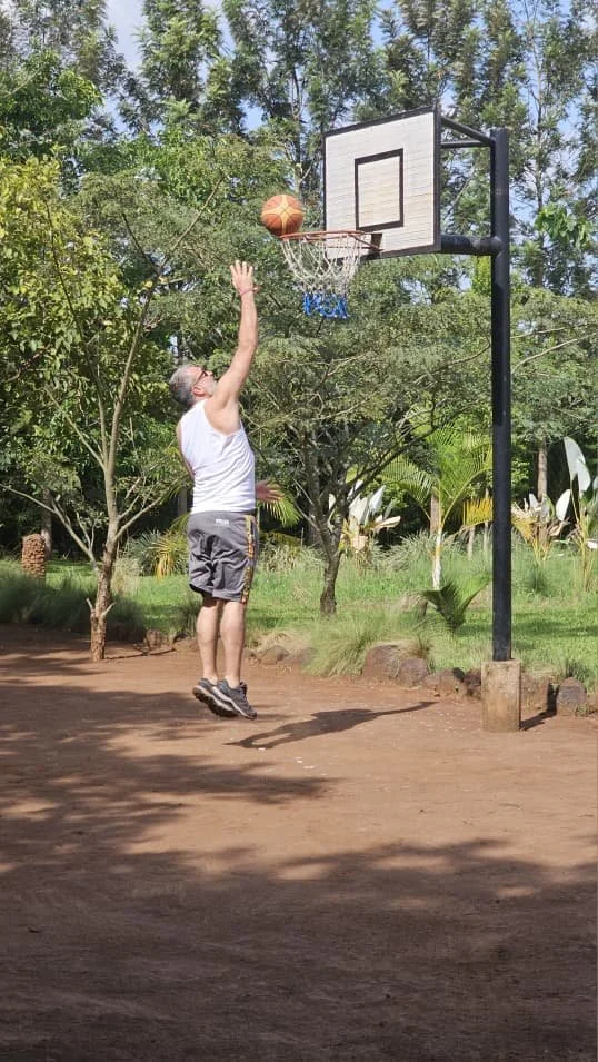 A man in white tank top and gray shorts jumping to make a basketball shot at an outdoor hoop in a park with trees and plants.