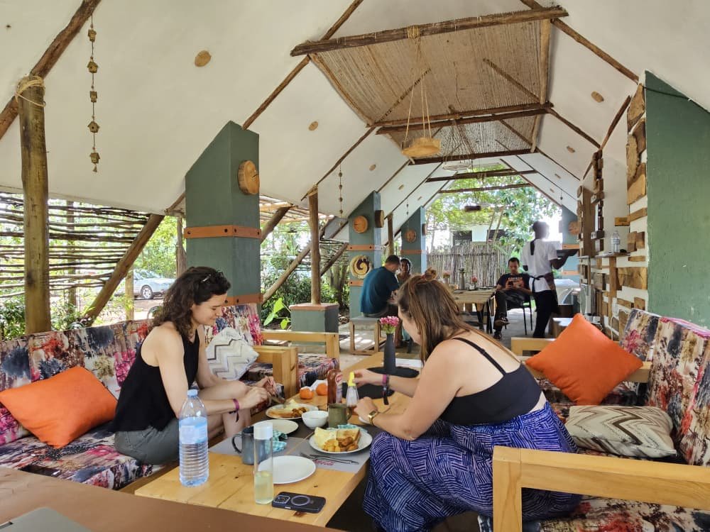 People dining in a rustic, covered outdoor restaurant with colorful sofas, pillows, and wooden décor.