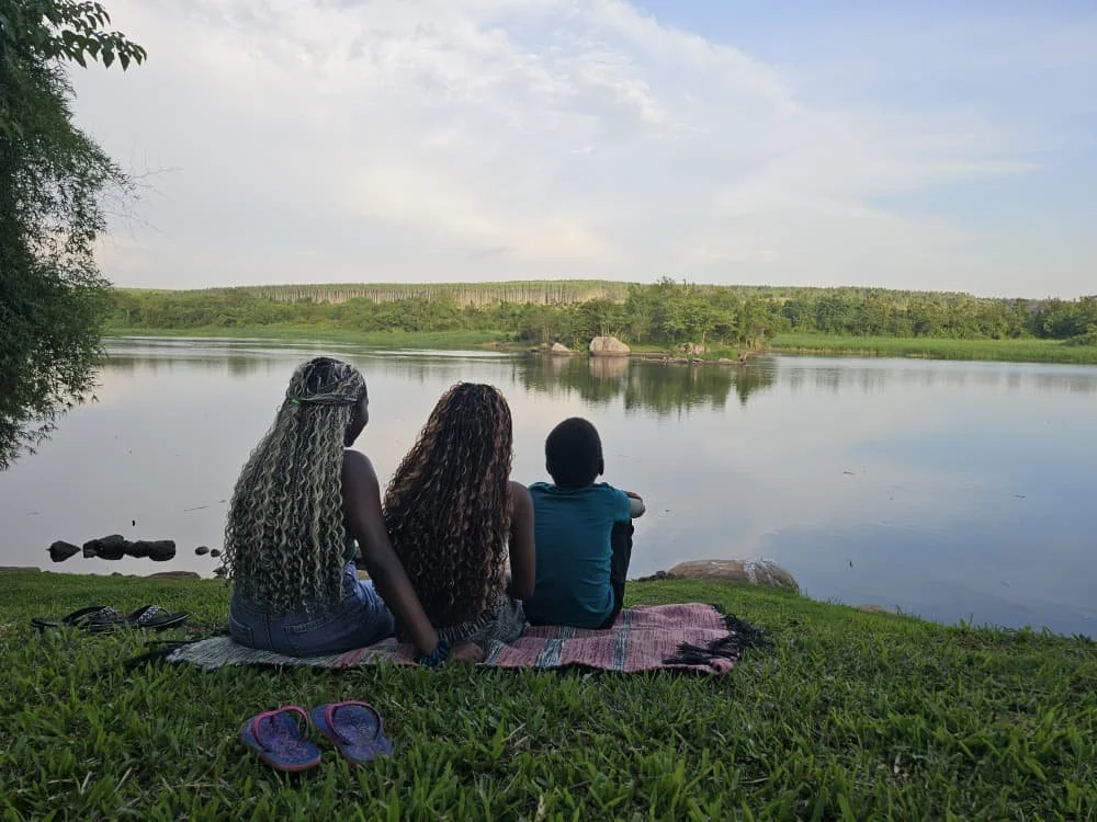Three children sitting on a blanket by a river, watching the water and greenery in the distance.