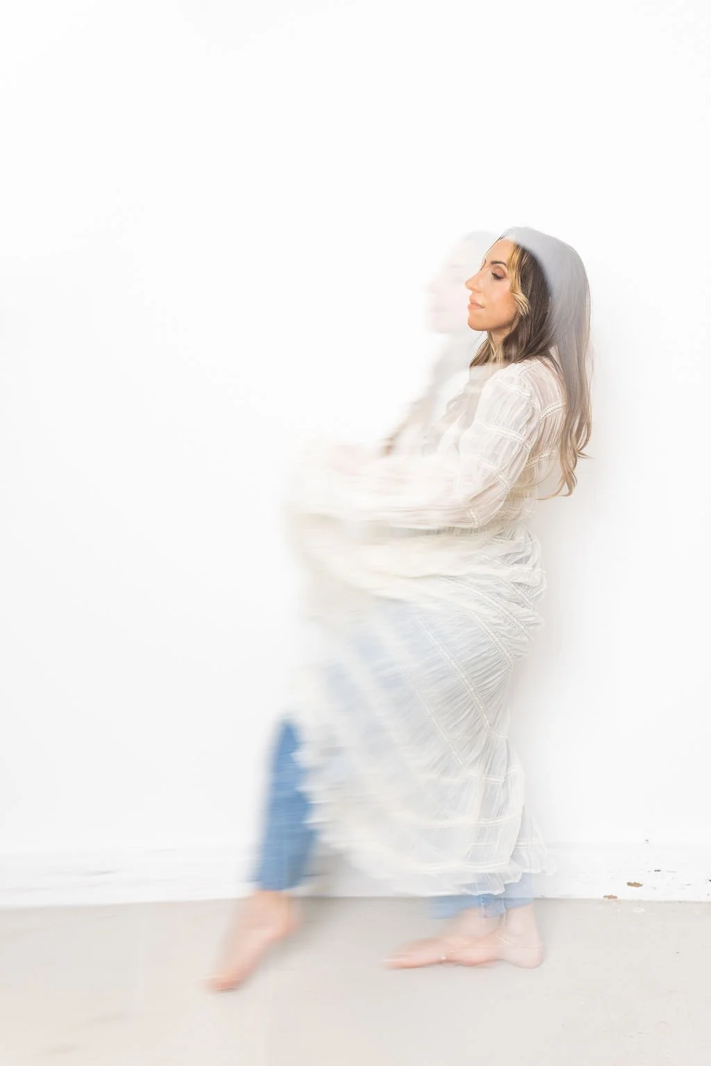 A woman in a white dress appears blurry, creating a ghostly effect as she stands leaning against a white wall.
