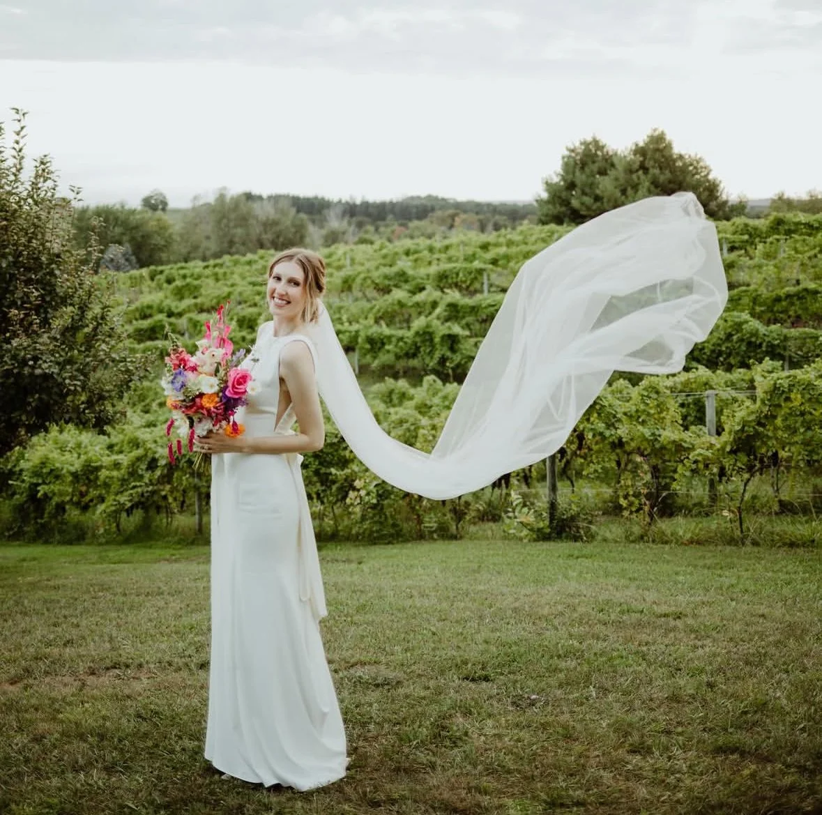 Bride stands in a vineyard holding a vibrant seasonal bouquet with pink, red, orange, blue & white flowers.