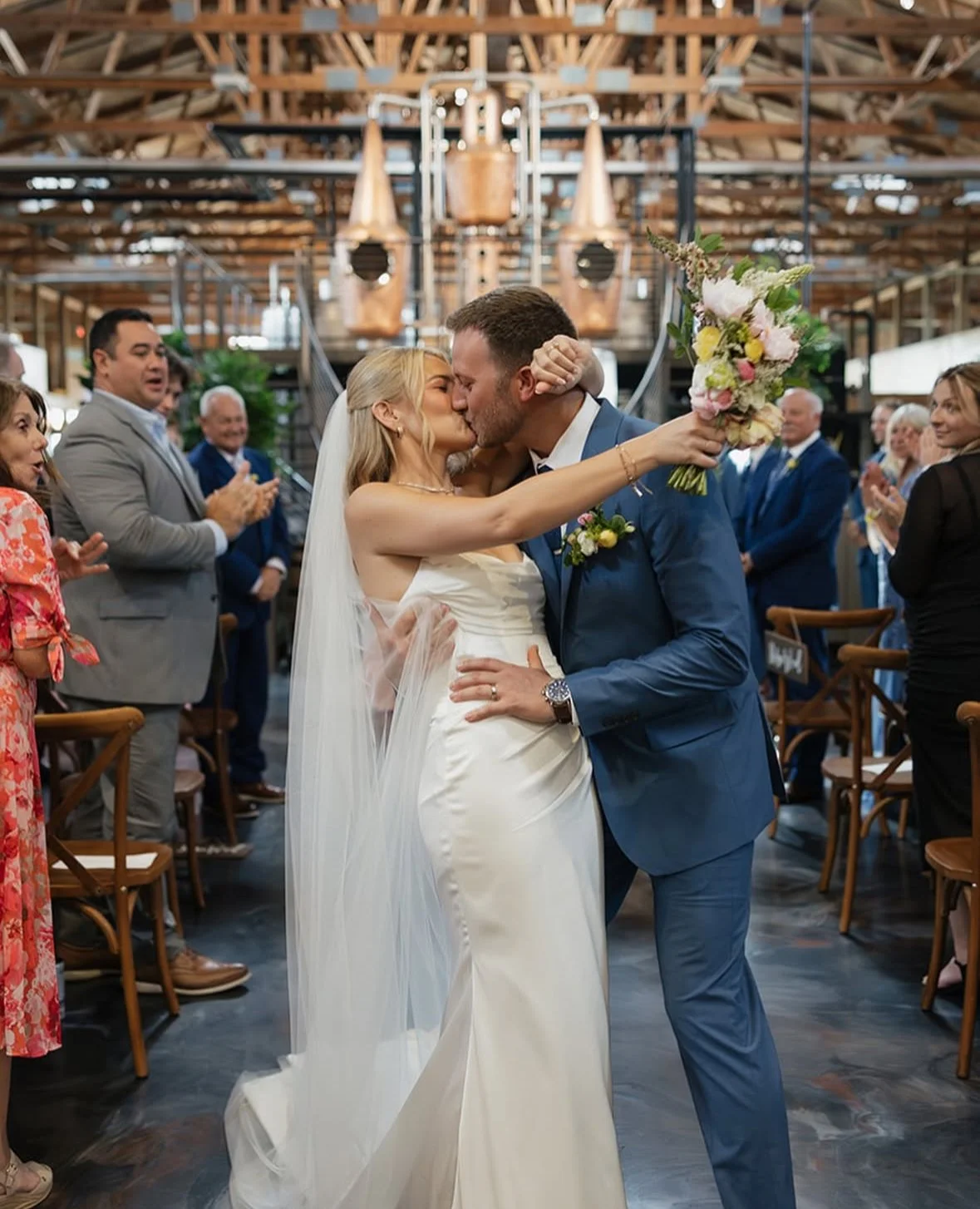 Taylor and Dillan kiss during their Gypsy Distillery wedding ceremony as Taylor holds a soft seasonal bouquet with white, blush, yellow & pink flowers.