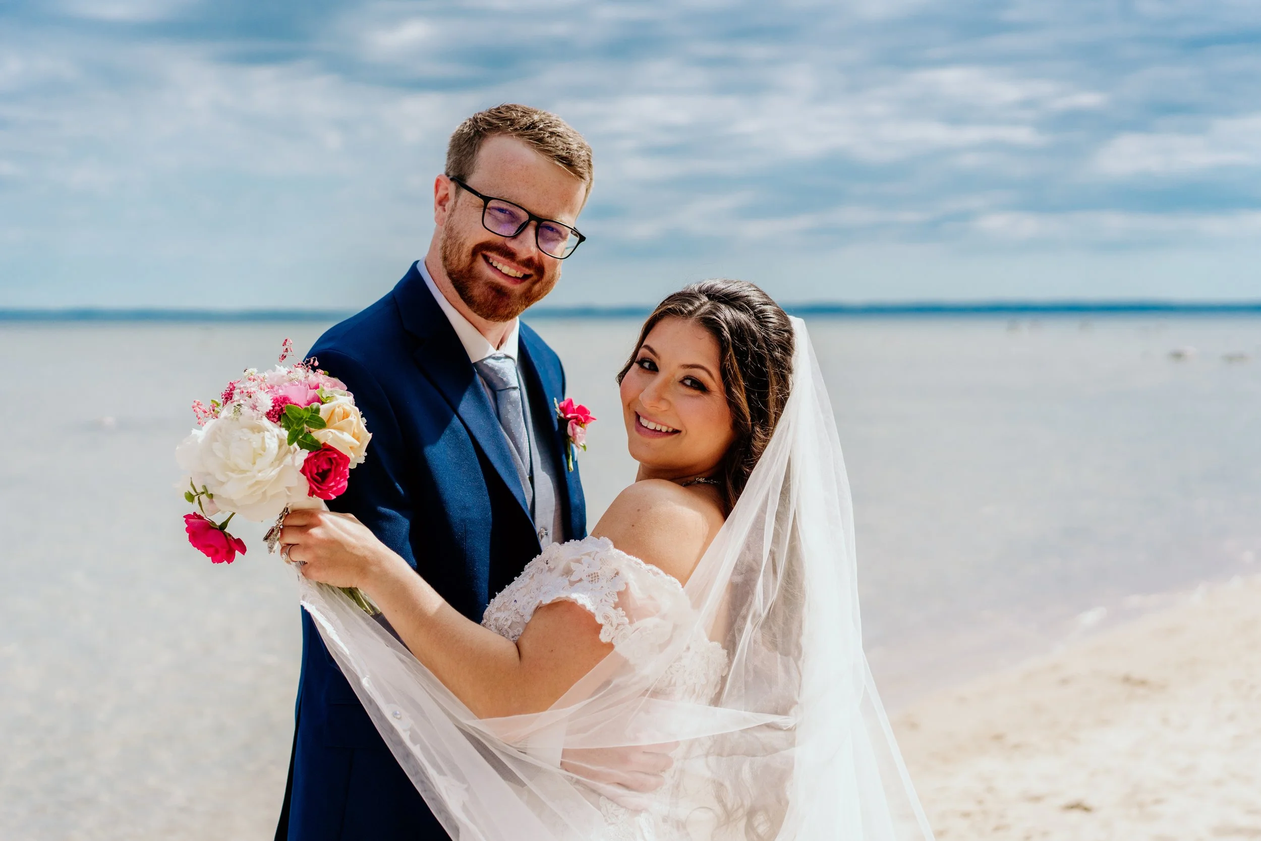 Newlyweds pose by the water as the bride holds a colorful wedding bouquet with white, pink, coral & yellow seasonal flowers.