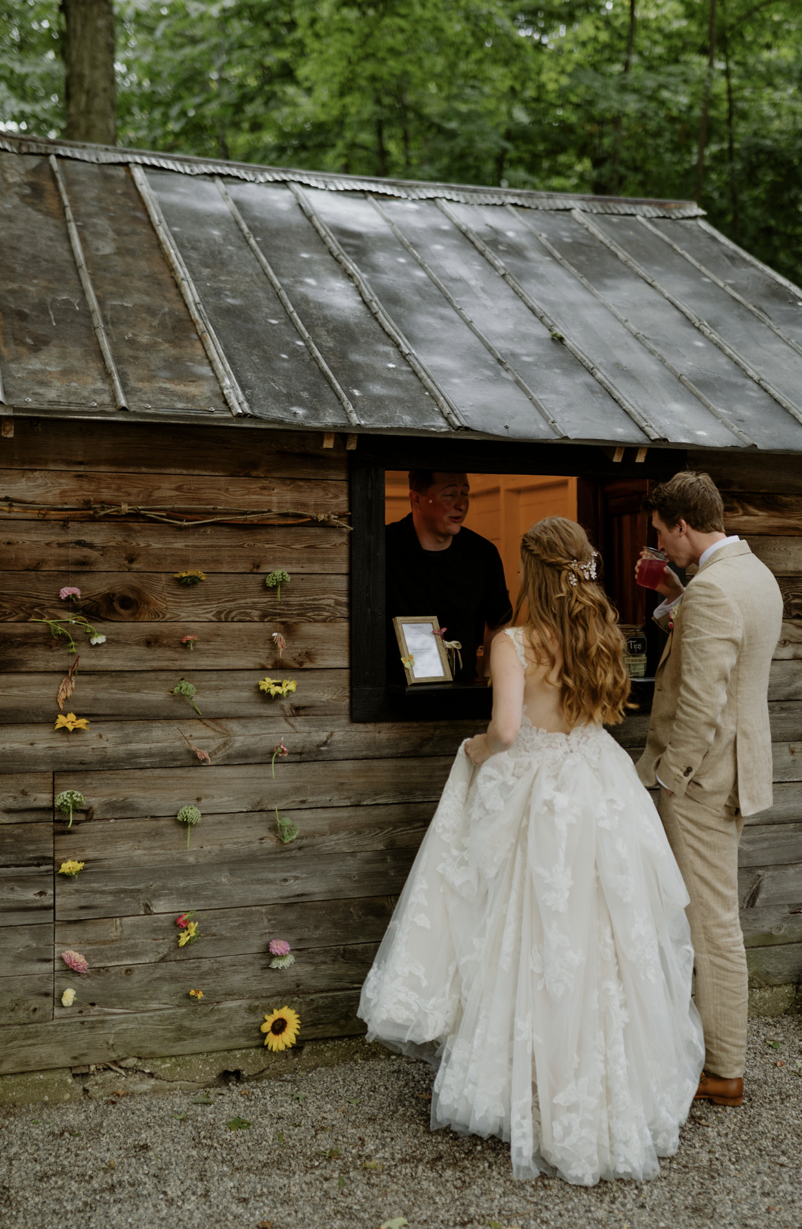 Bride and groom ordering drinks at rustic outdoor wedding bar with natural wood structure and subtle floral details