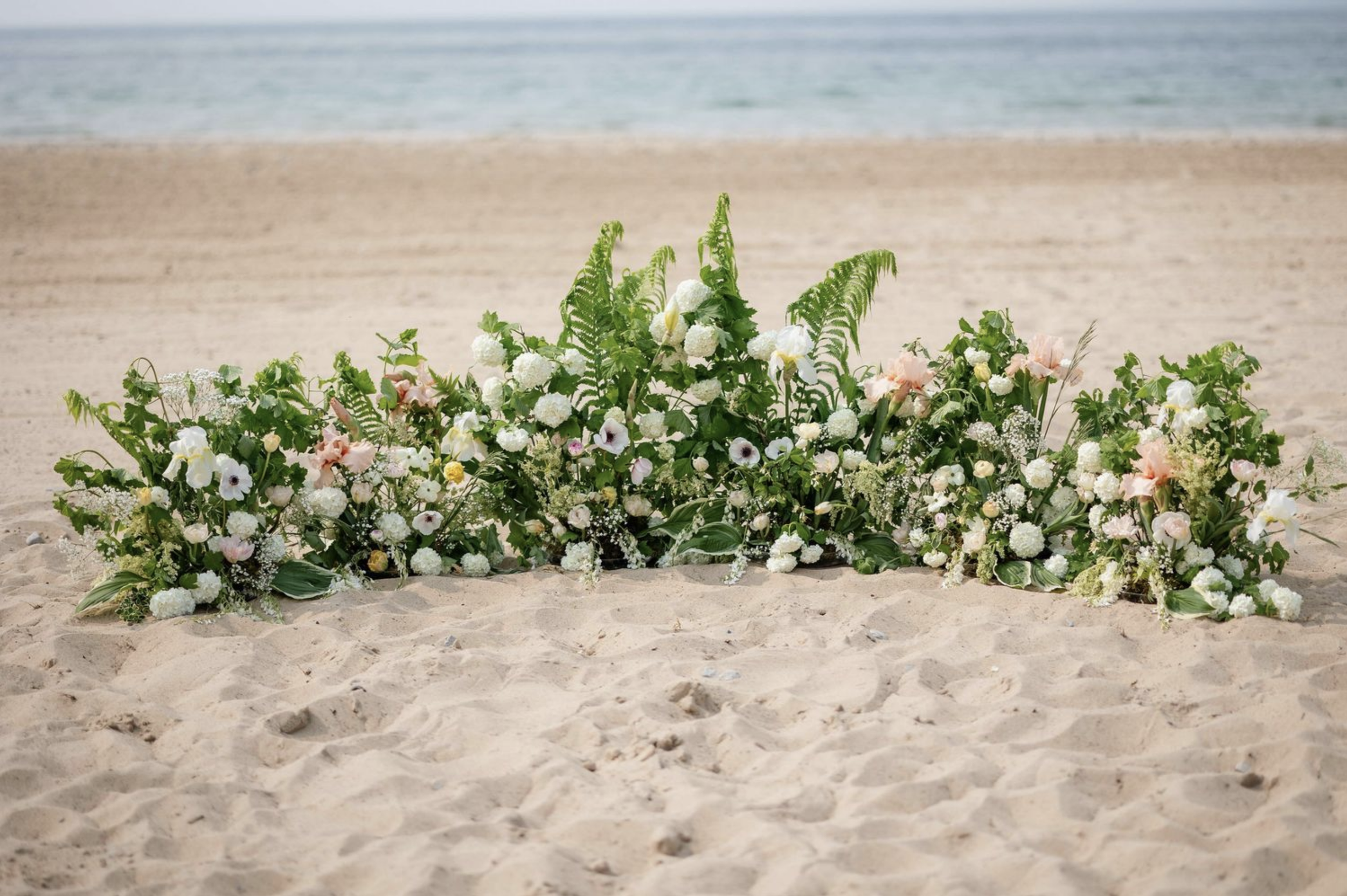 Beach wedding ceremony flowers arranged in a lush, low installation with white and soft pastel blooms along the sand