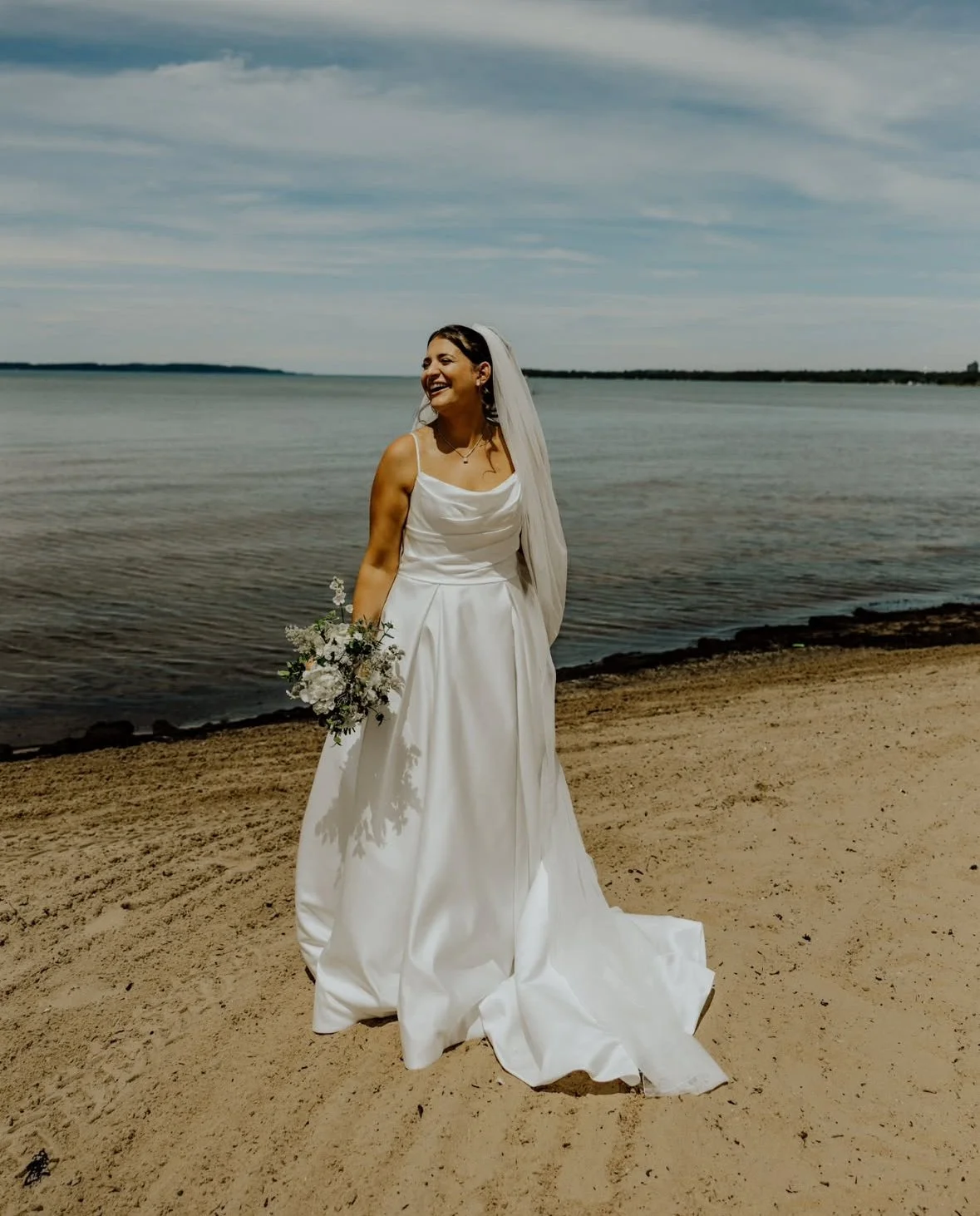 Katie stands on the beach at Harbor Bay Inn in her wedding dress, holding a soft white and green seasonal bridal bouquet beside the water.