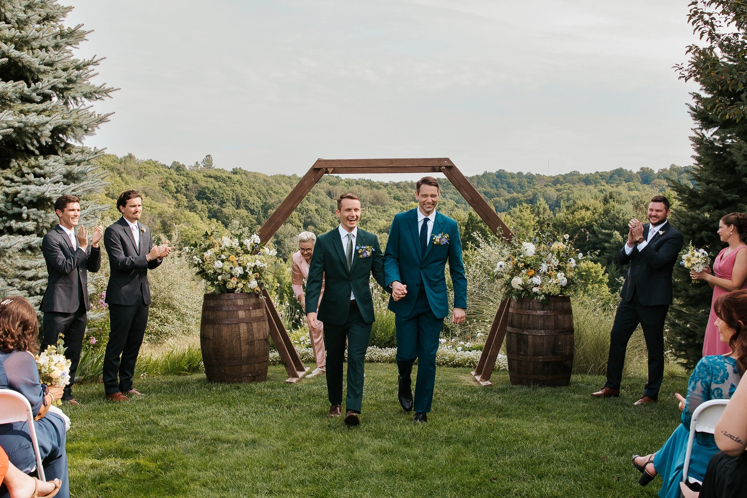 Newlyweds walk back down the aisle after an outdoor ceremony framed by colorful seasonal floral arrangements on wine barrels.