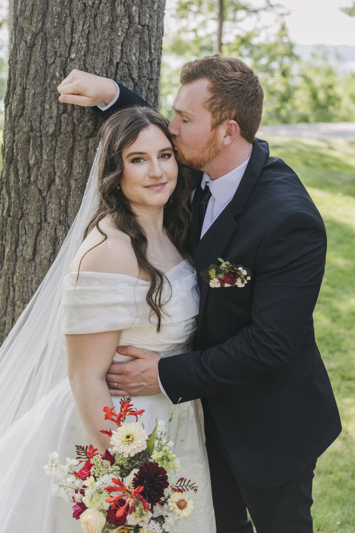Lillian and Ryan pose together outdoors on their wedding day, with Lillian holding a colorful seasonal bouquet in red, burgundy, cream & green tones.