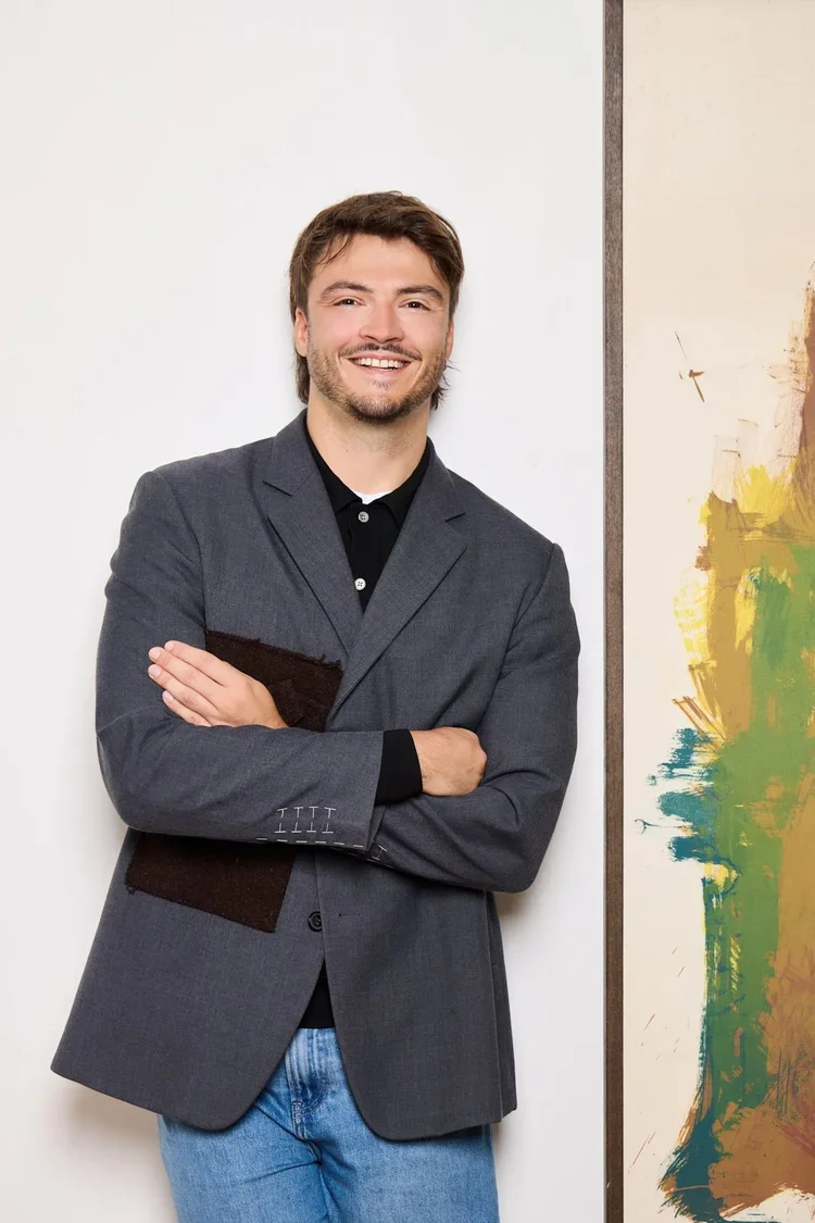 Jeune homme souriant portant une veste grise, tenant un document ou un livre, debout à côté d'une œuvre d'art abstraite avec des couleurs vives.