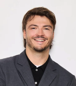 Homme souriant avec barbe, portant un costume gris, fond blanc