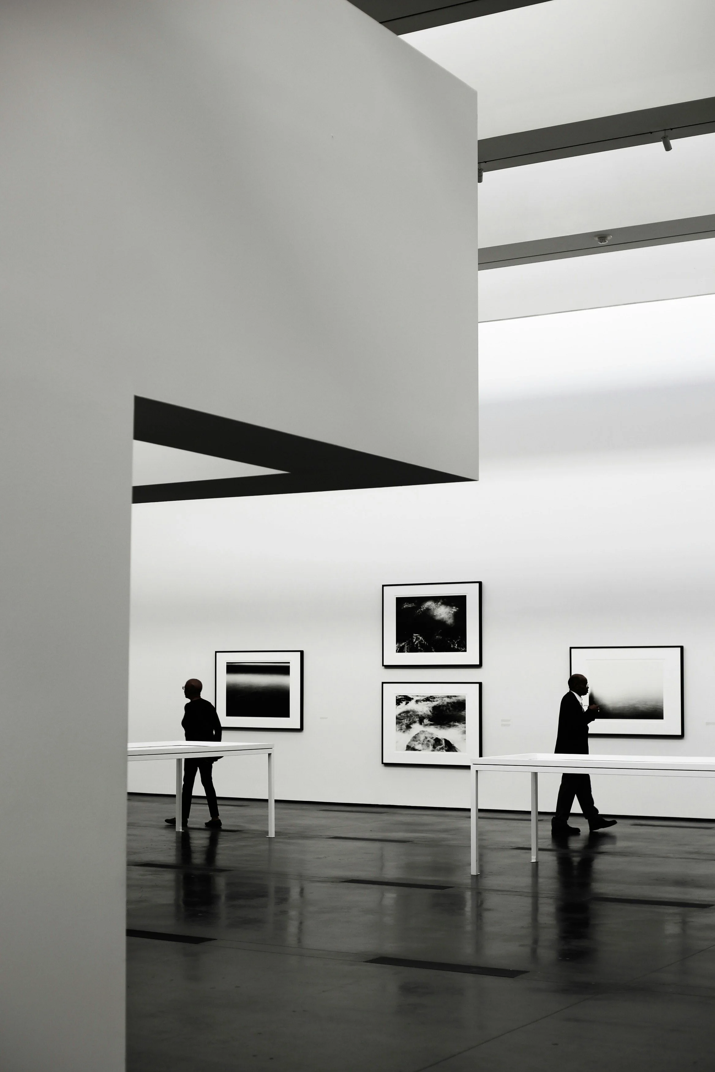 Vue d'une galerie d'art avec plusieurs œuvres photographiques en noir et blanc accrochées au mur. Trois personnes sont visibles, deux debout et un en mouvement, dans un espace lumineux avec plafond blanc.