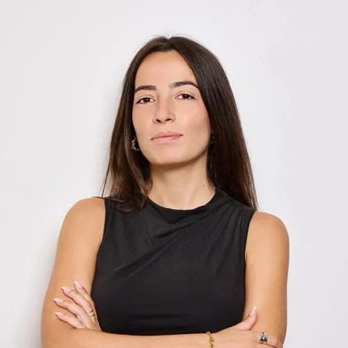 Une femme avec des cheveux longs et roux, portant un haut noir sans manches, avec les bras croisés, se tenant contre un fond blanc.