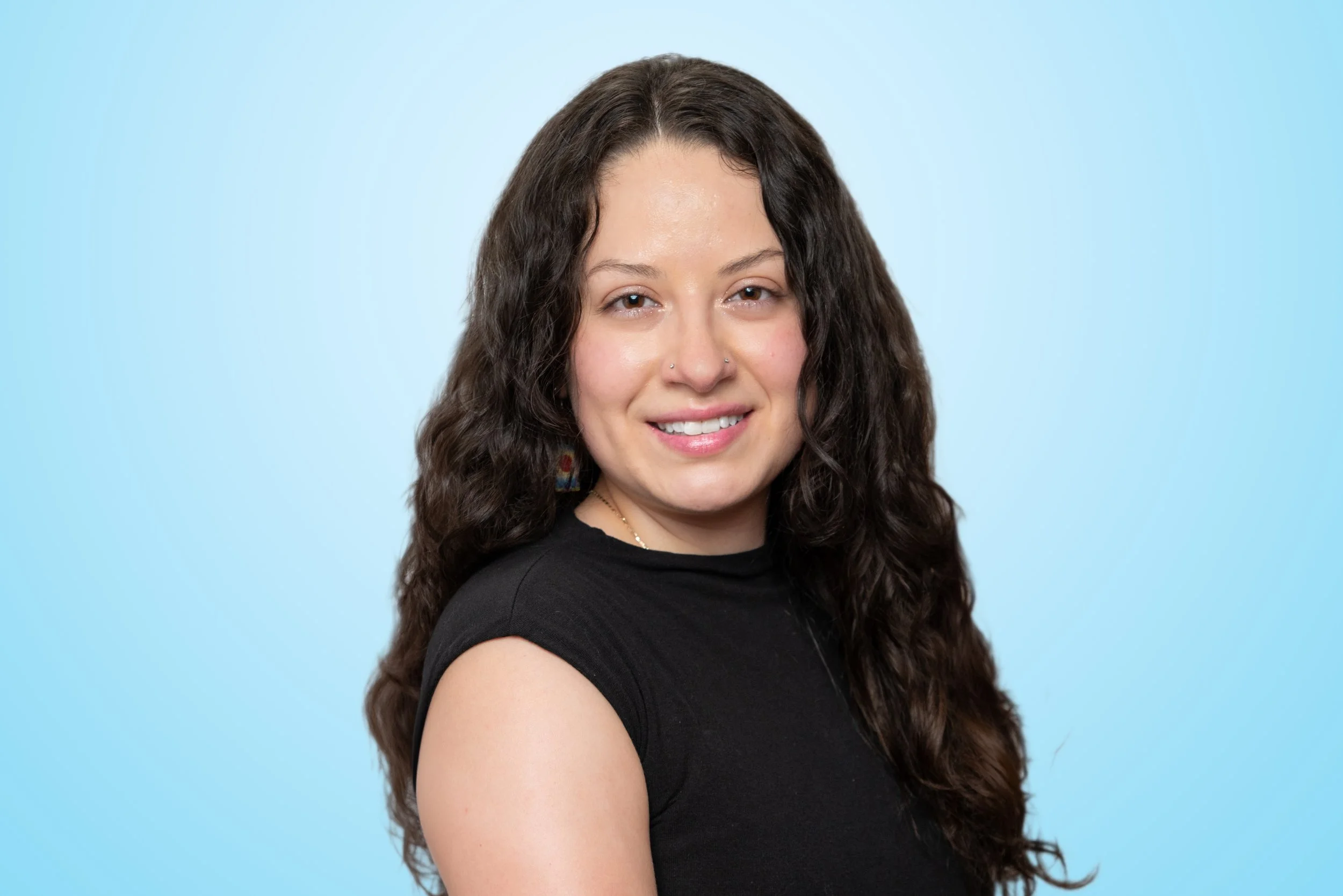 Portrait of a young woman with long curly hair, smiling, wearing a black top, against a light blue background.