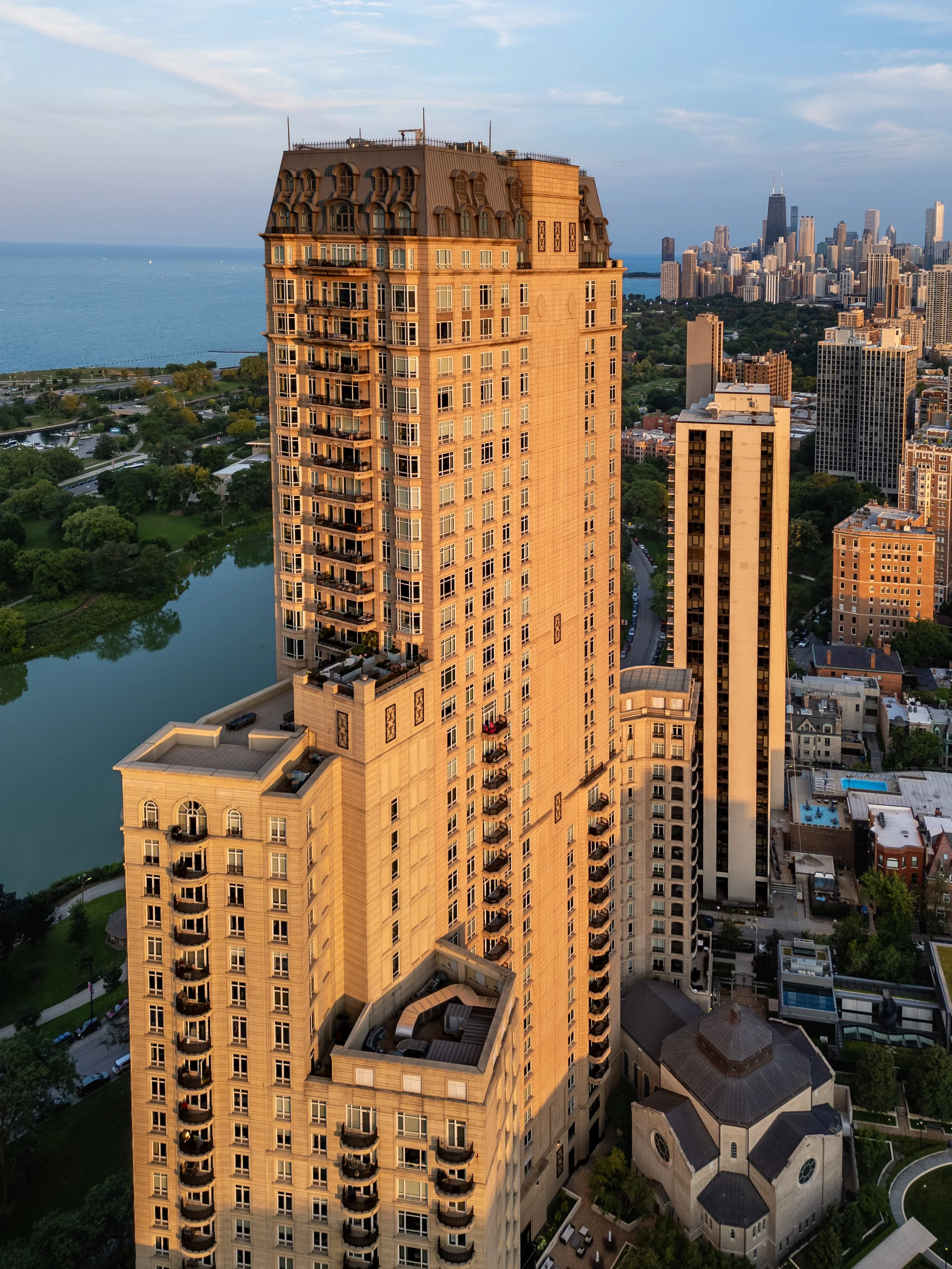 A tall beige high-rise building with multiple balconies and a detailed rooftop, situated near a river and city skyline in the distance, during sunset.