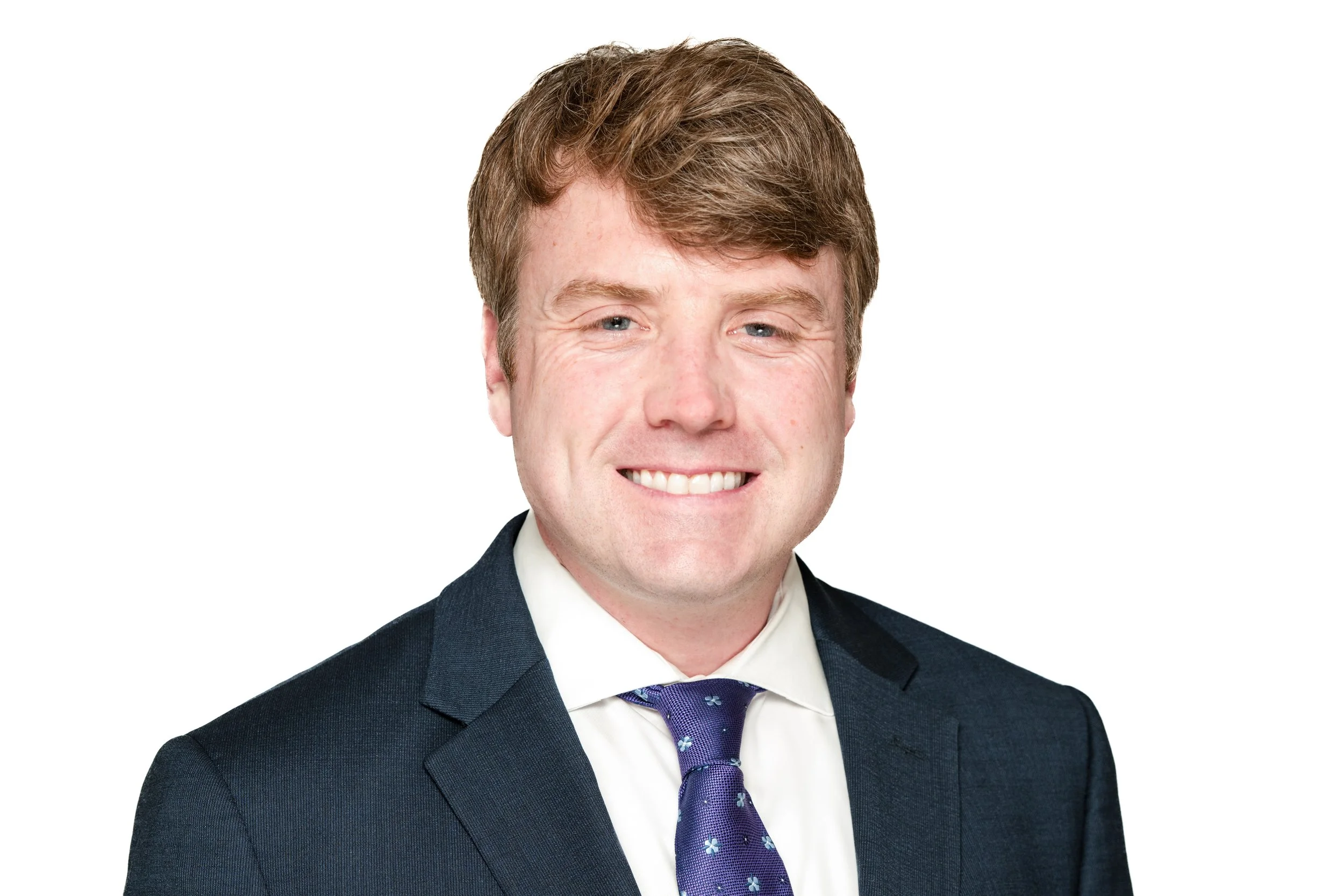 Portrait of a smiling man with brown hair wearing a dark suit, white shirt, and purple tie against a white background.