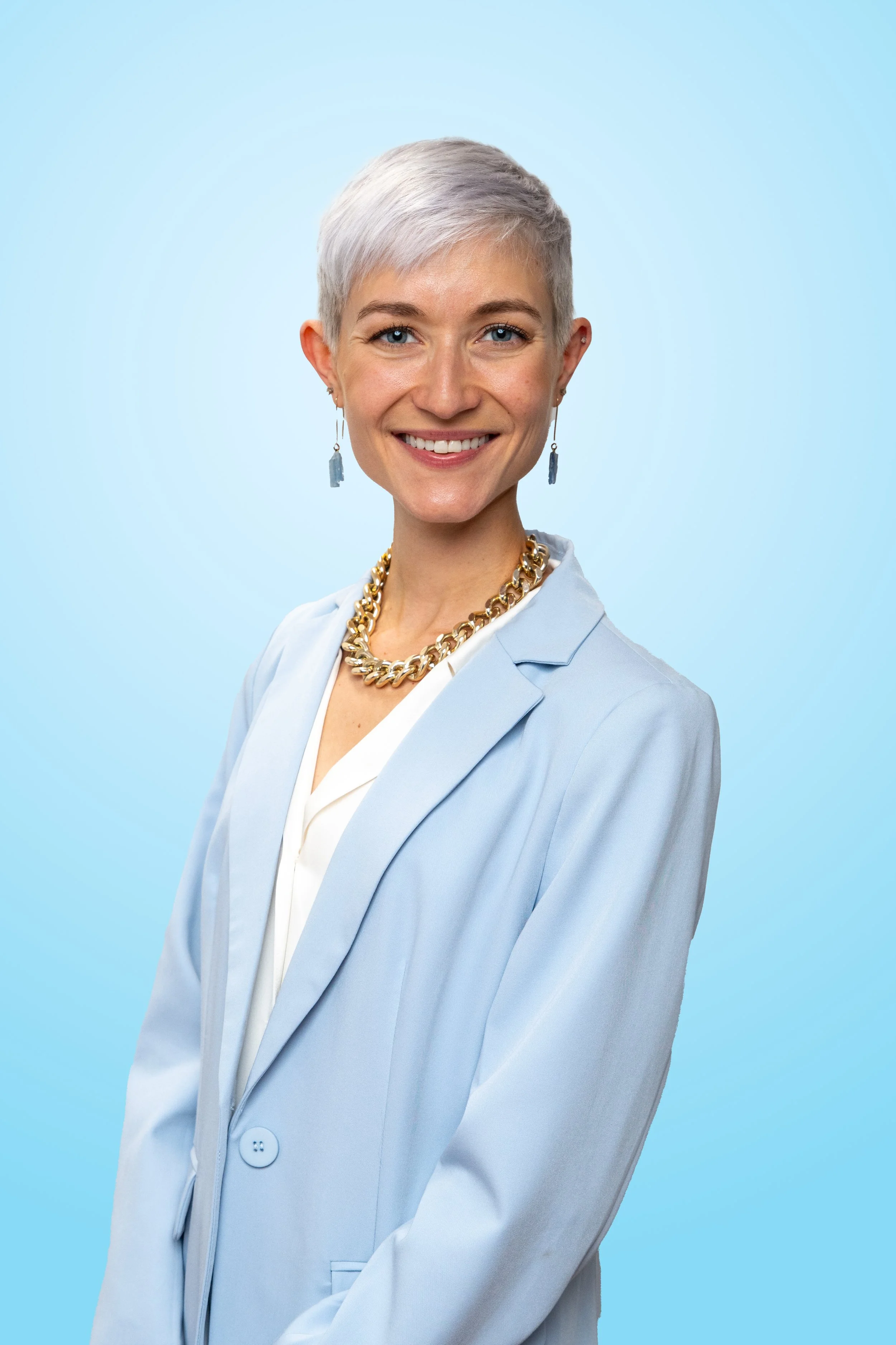 A woman with short platinum blonde hair, wearing a light blue blazer, white top, gold chain necklace, and matching earrings, standing against a light blue background and smiling at the camera.