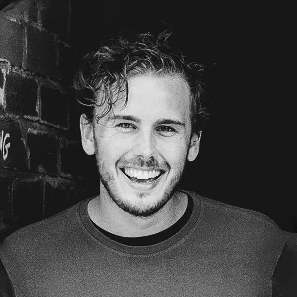 Black and white photo of a young man with curly hair, smiling and wearing a t-shirt, standing against a brick wall.