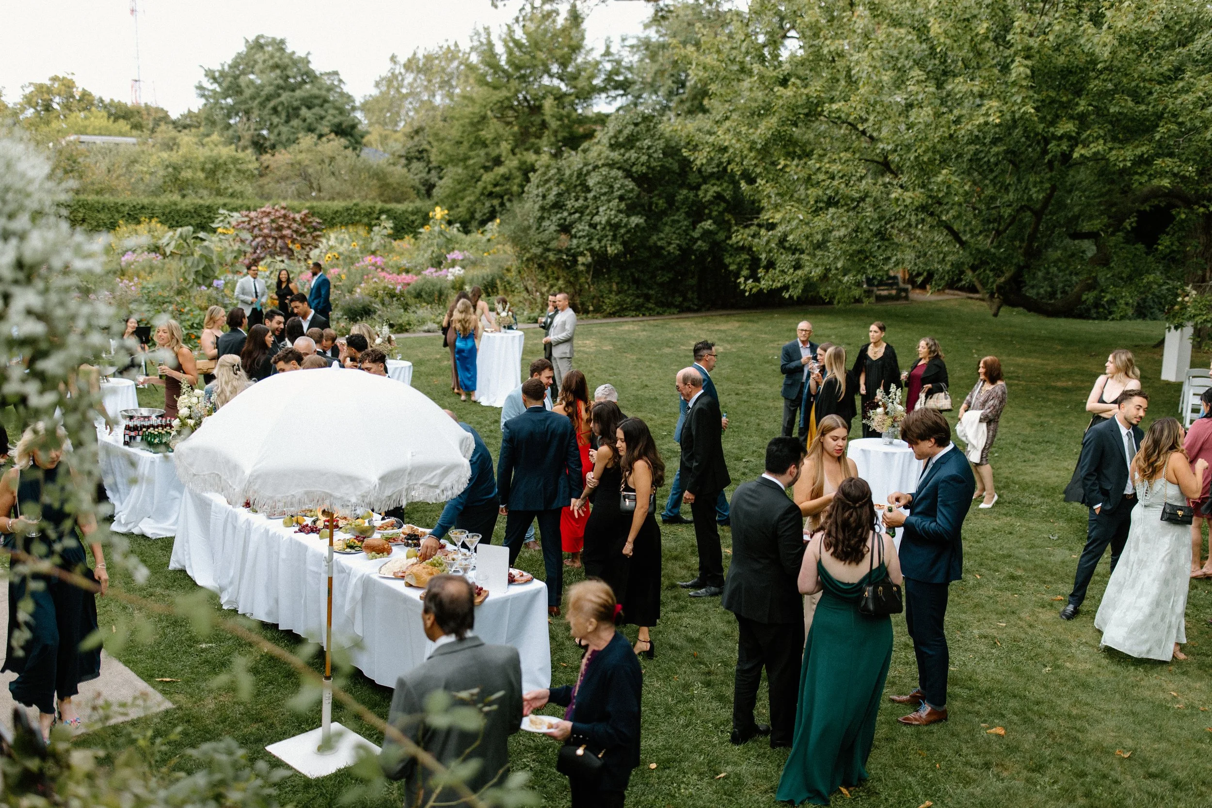 People attending an outdoor garden reception with tables of food, drink, and floral arrangements on a lush green lawn surrounded by trees and flowering bushes.