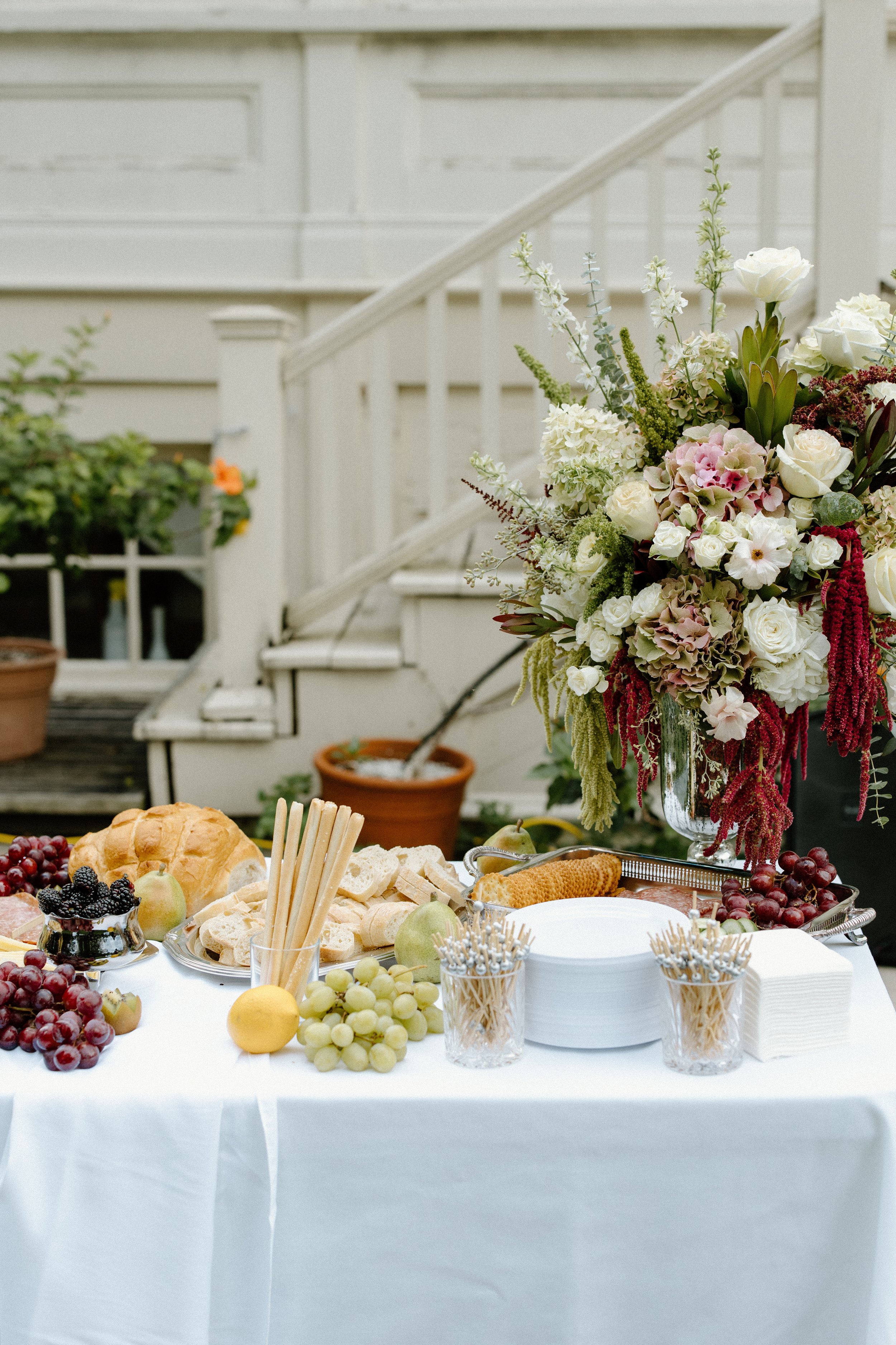 A table set for a wedding reception with a white tablecloth, assorted bread, grapes, apples, and a large floral arrangement with white, pink, and red flowers in a glass vase.