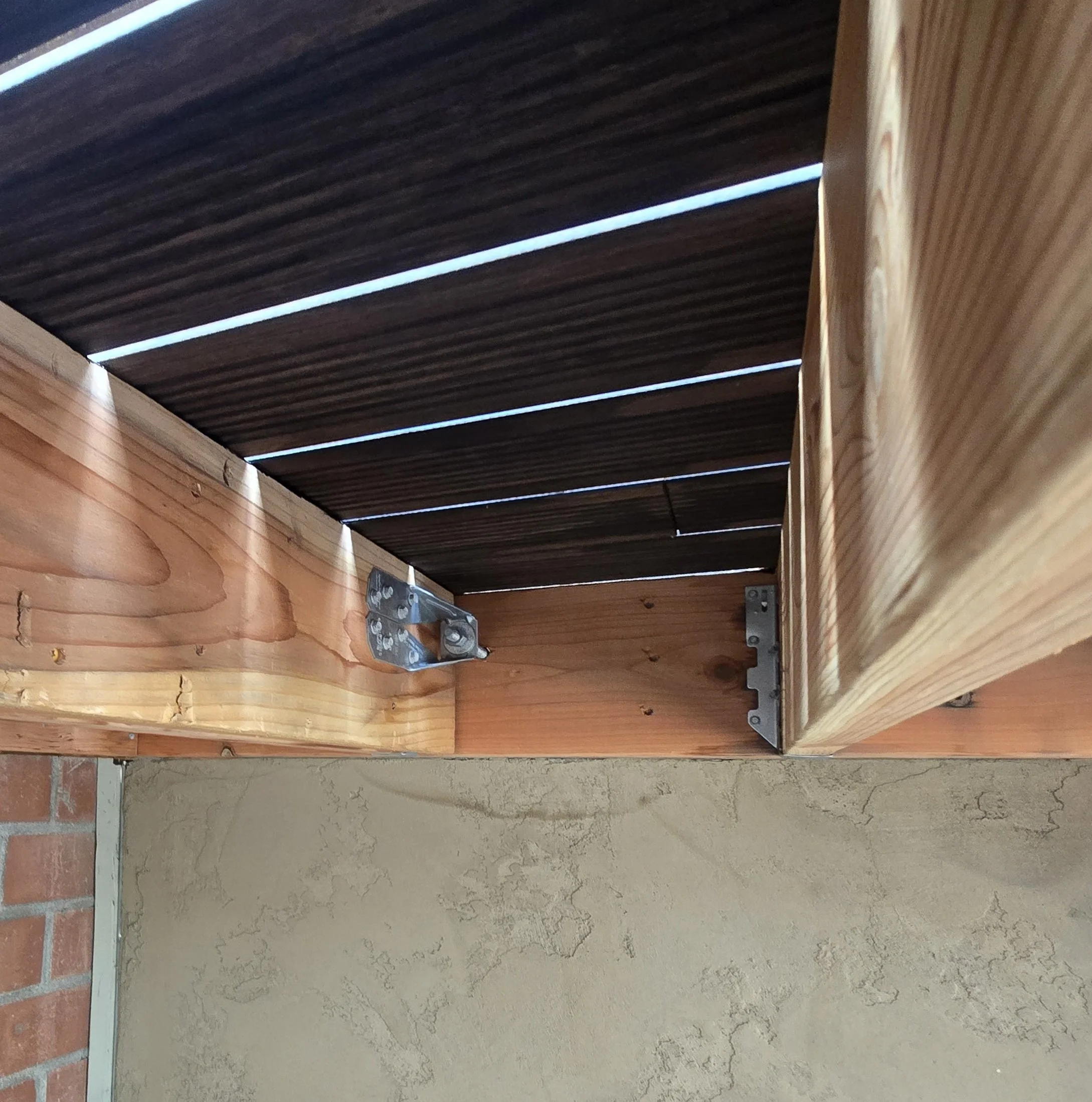 A underside view of a redwood deck frame showing light-colored joists secured to a beam with galvanized steel joist hangers and angle brackets.