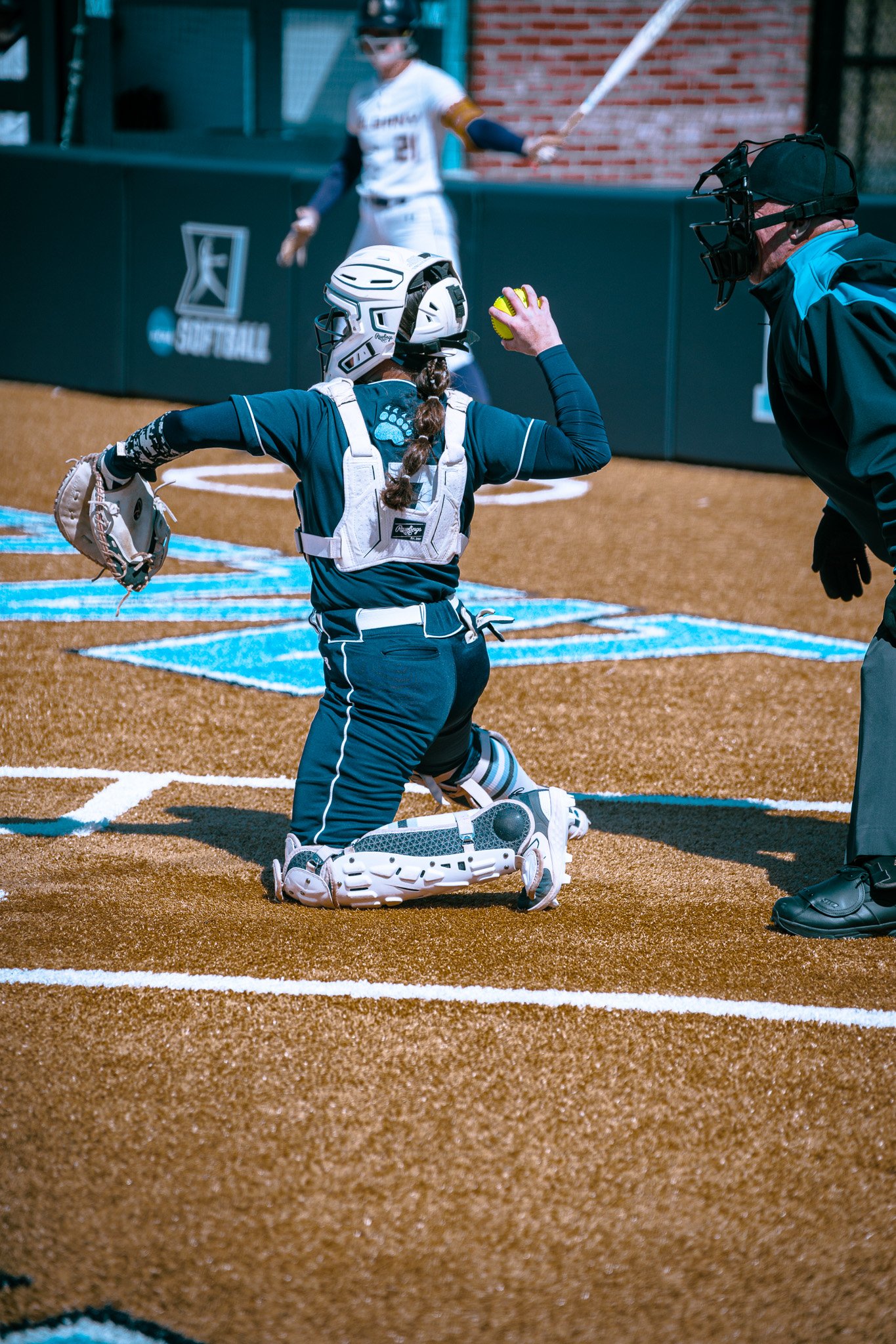 Softball catcher preparing to throw ball, umpire nearby, and batter in the background.