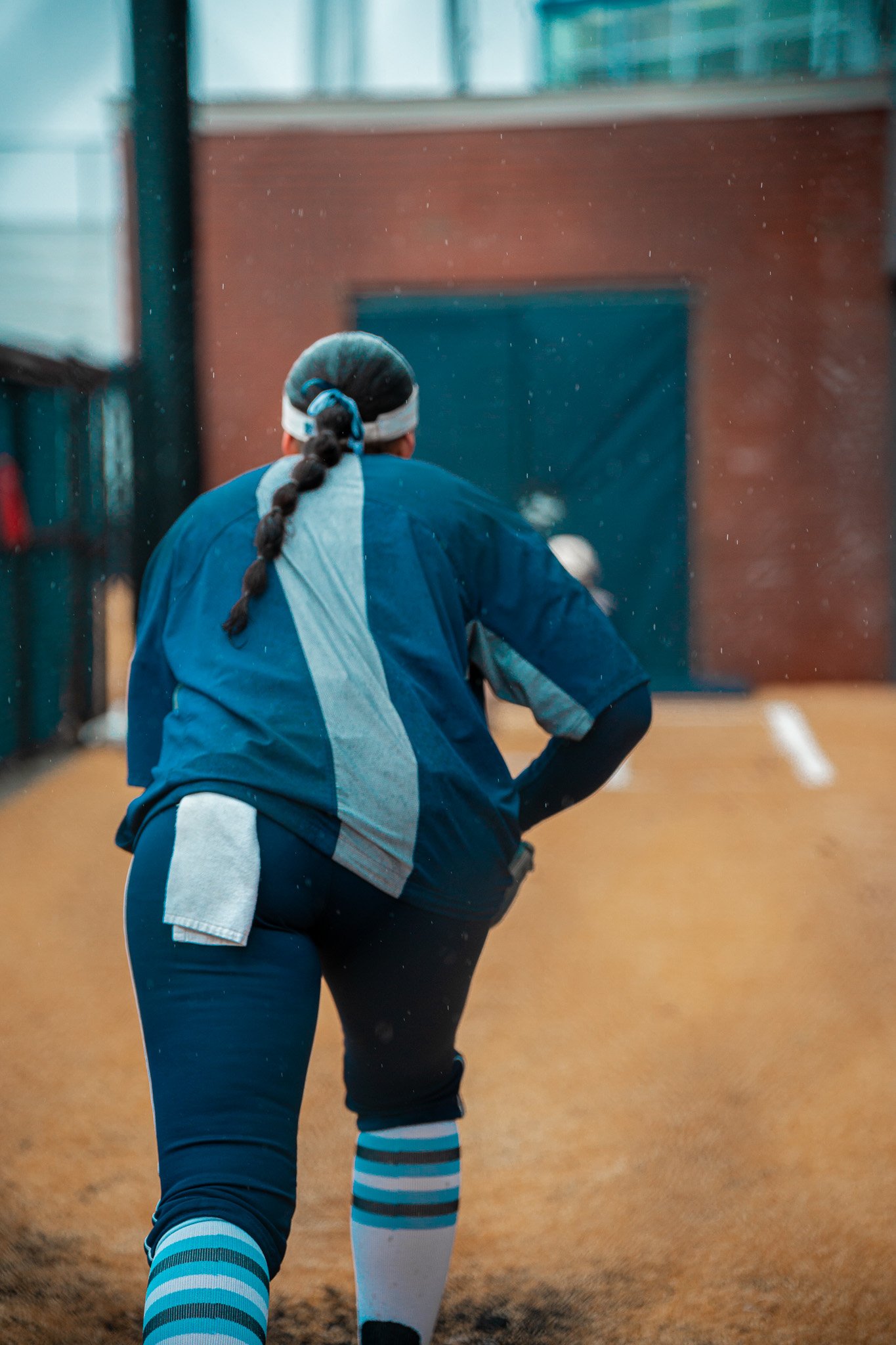 Softball player with braided hair, in blue uniform, preparing to pitch on a dirt field.
