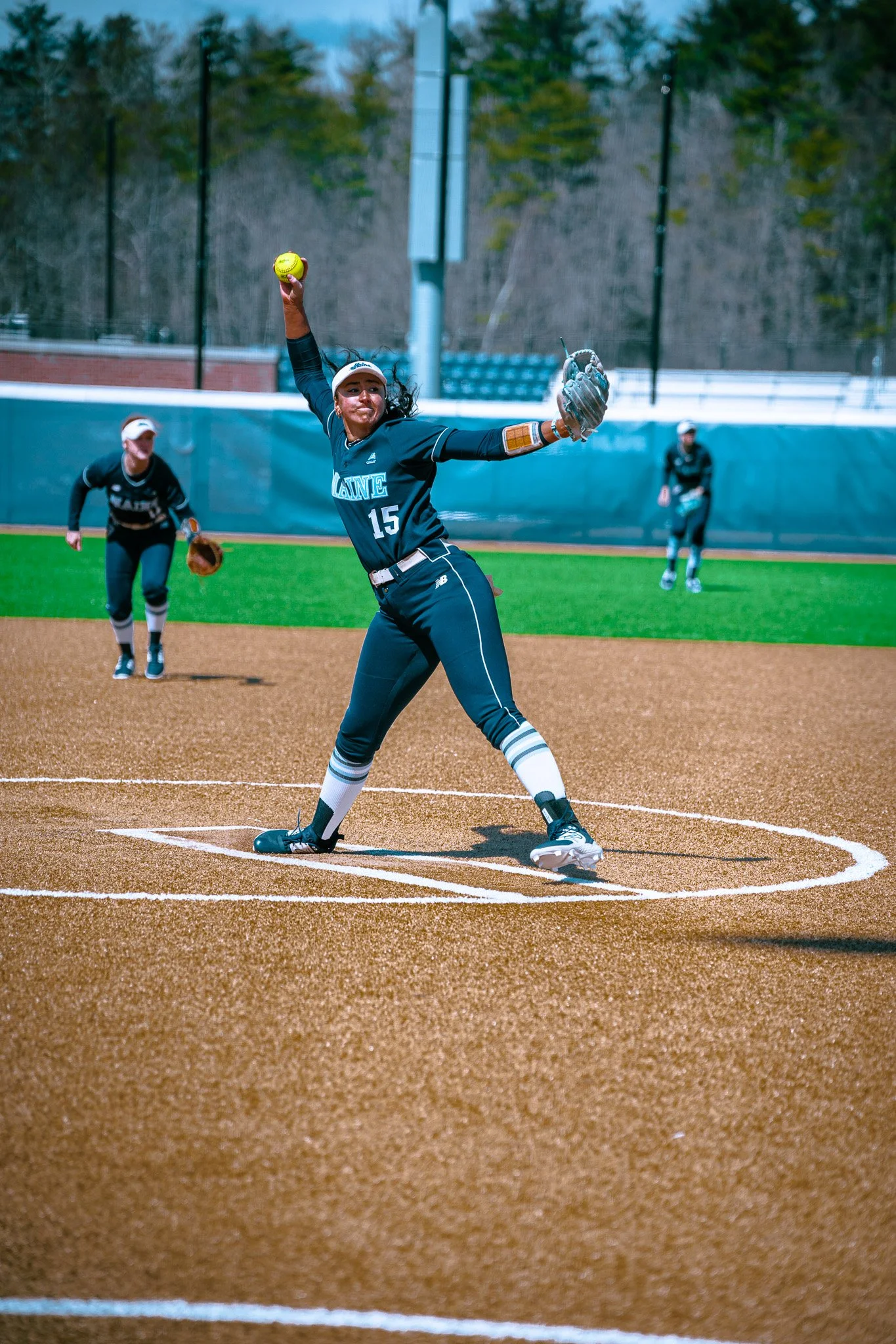 Softball pitcher in action during a game on a baseball field, wearing a blue uniform with the text "MAINE" and the number 15.