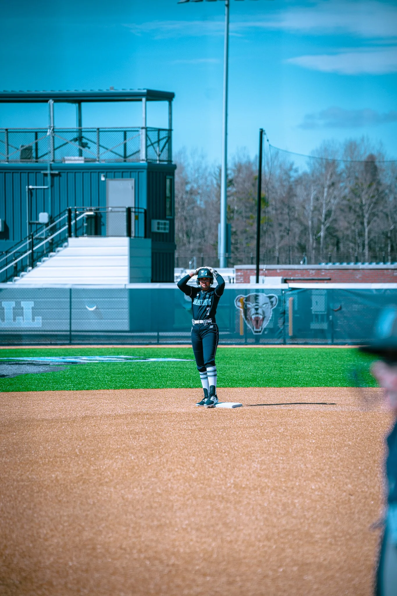 Baseball player standing on second base with hands on the helmet, outdoor field, blue building in background.