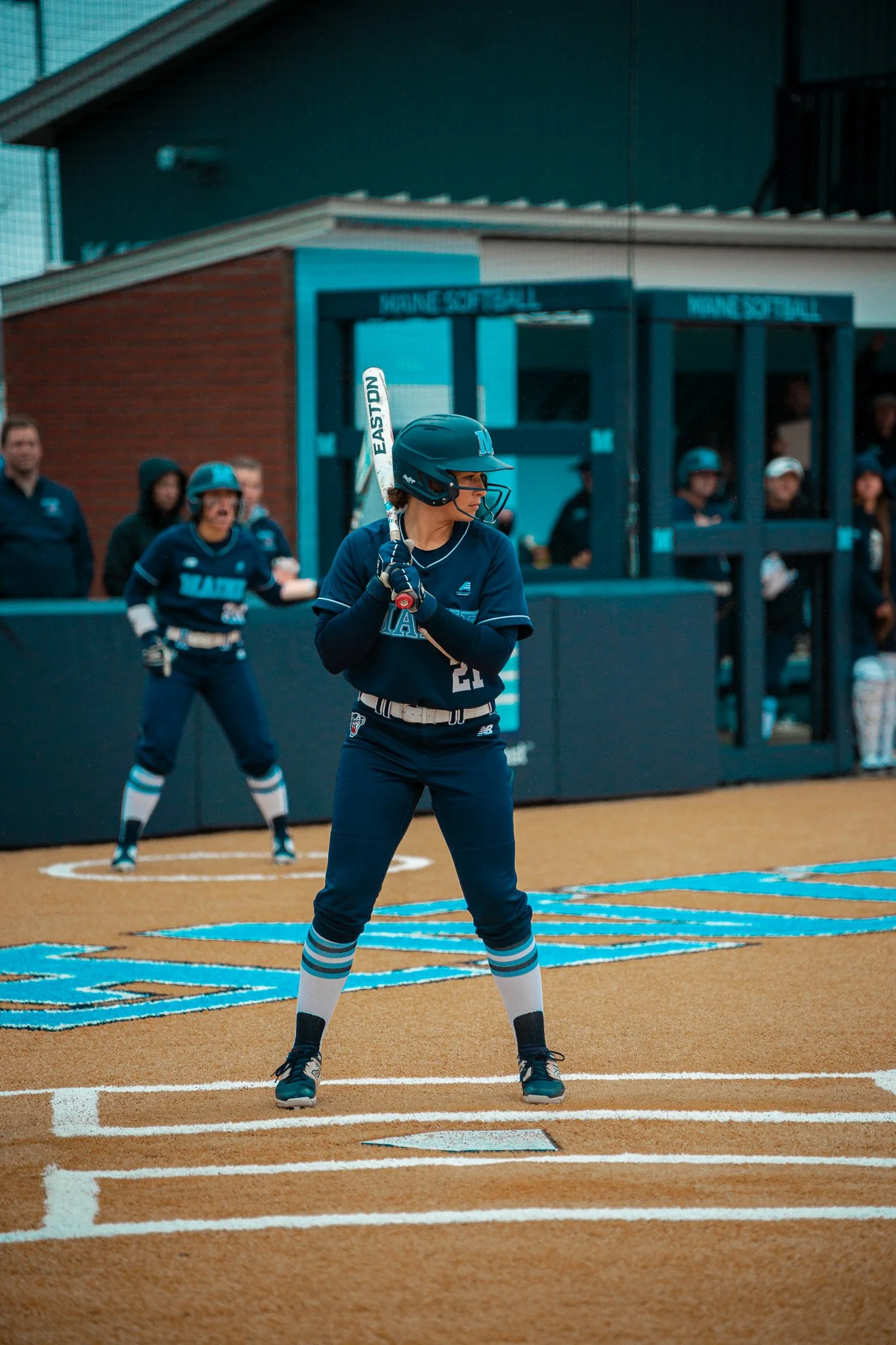 Softball player in batting stance during a game at Maine softball field, wearing a helmet and uniform. Teammates and spectators visible in the background.