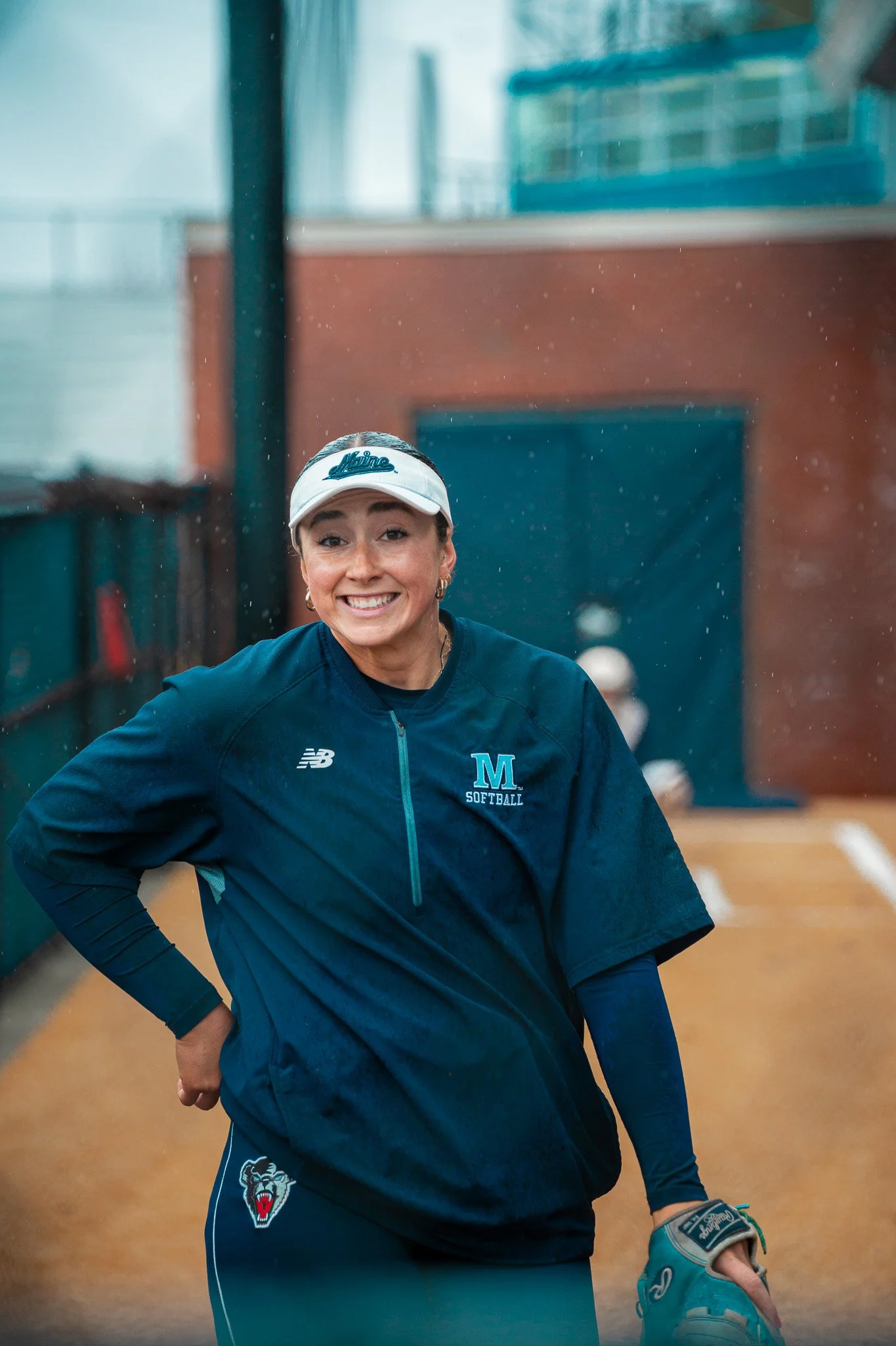 A smiling person wearing a blue softball uniform and white visor with "Maine" written on it, standing on a sports field under a cloudy sky. They are holding a softball glove.