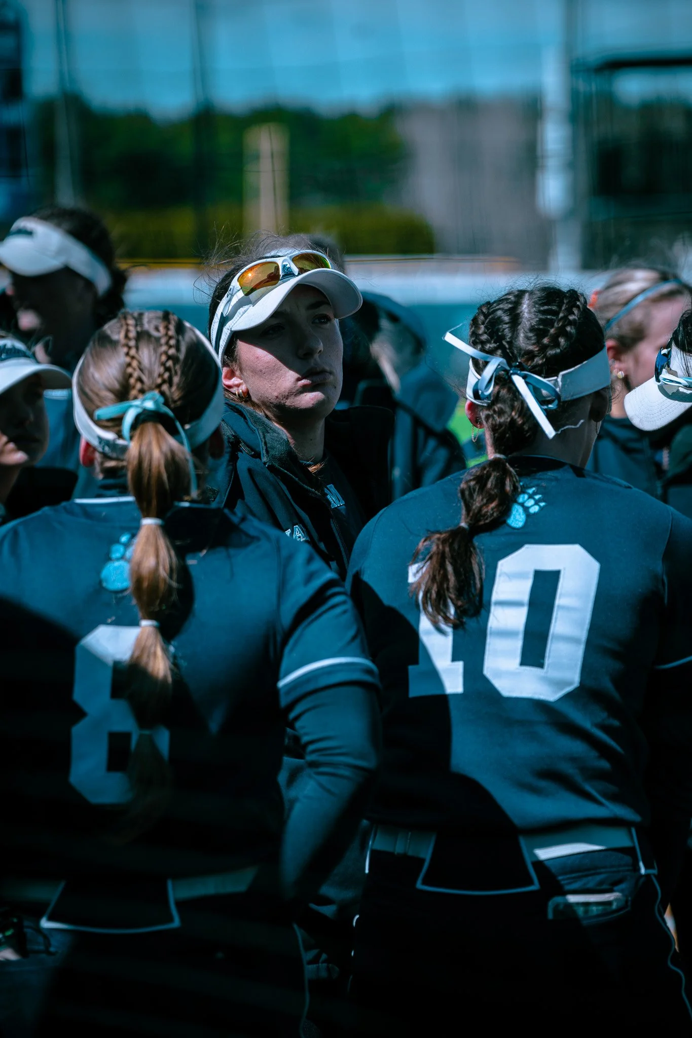 Softball team in a huddle, wearing uniforms and visors, with numbers 8 and 10 visible.