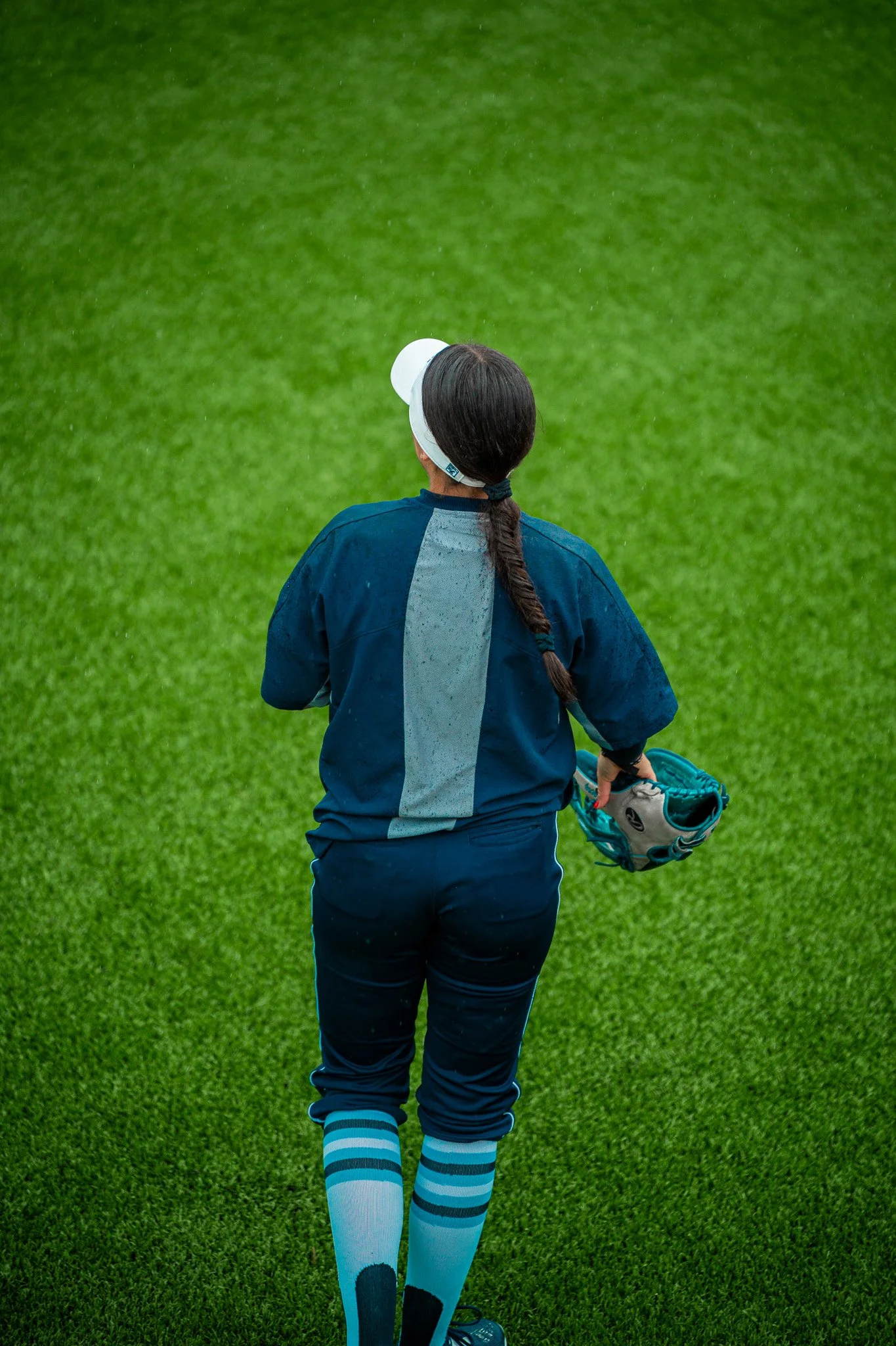 Softball player wearing blue uniform holding a glove on a grass field.