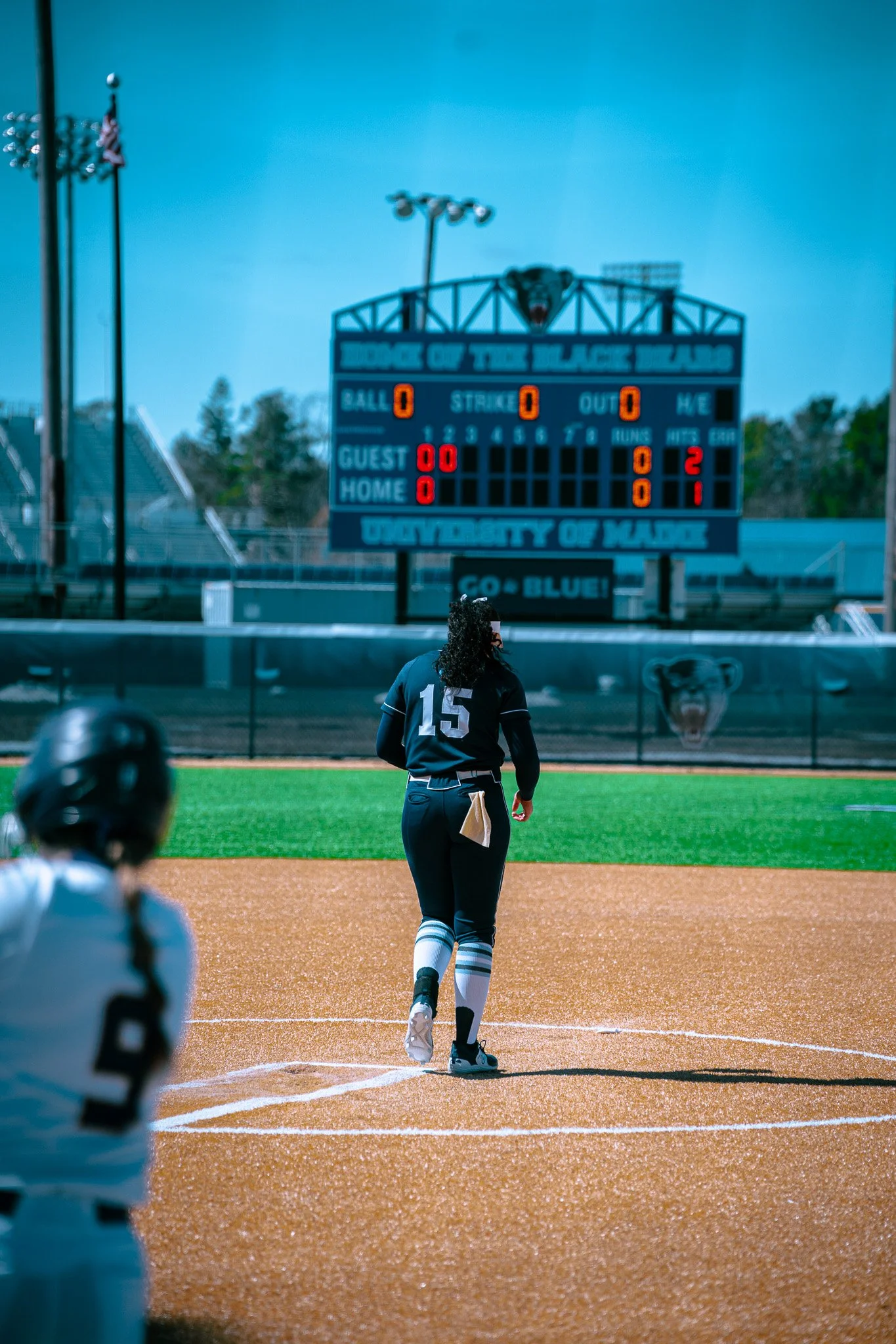 Softball players on a field with a scoreboard in the background displaying balls, strikes, and outs, at the University of Maine.