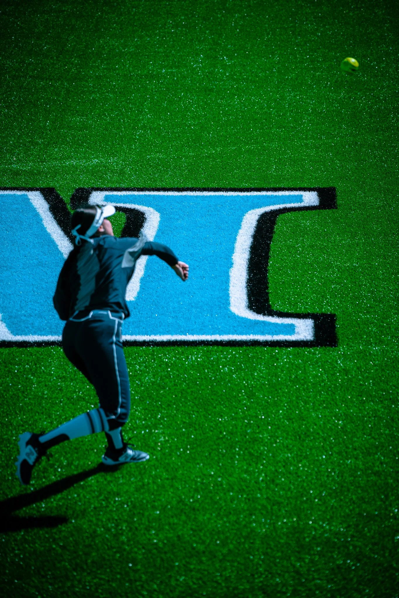Softball player pitching on a green field with marked lines, wearing a visor and uniform.