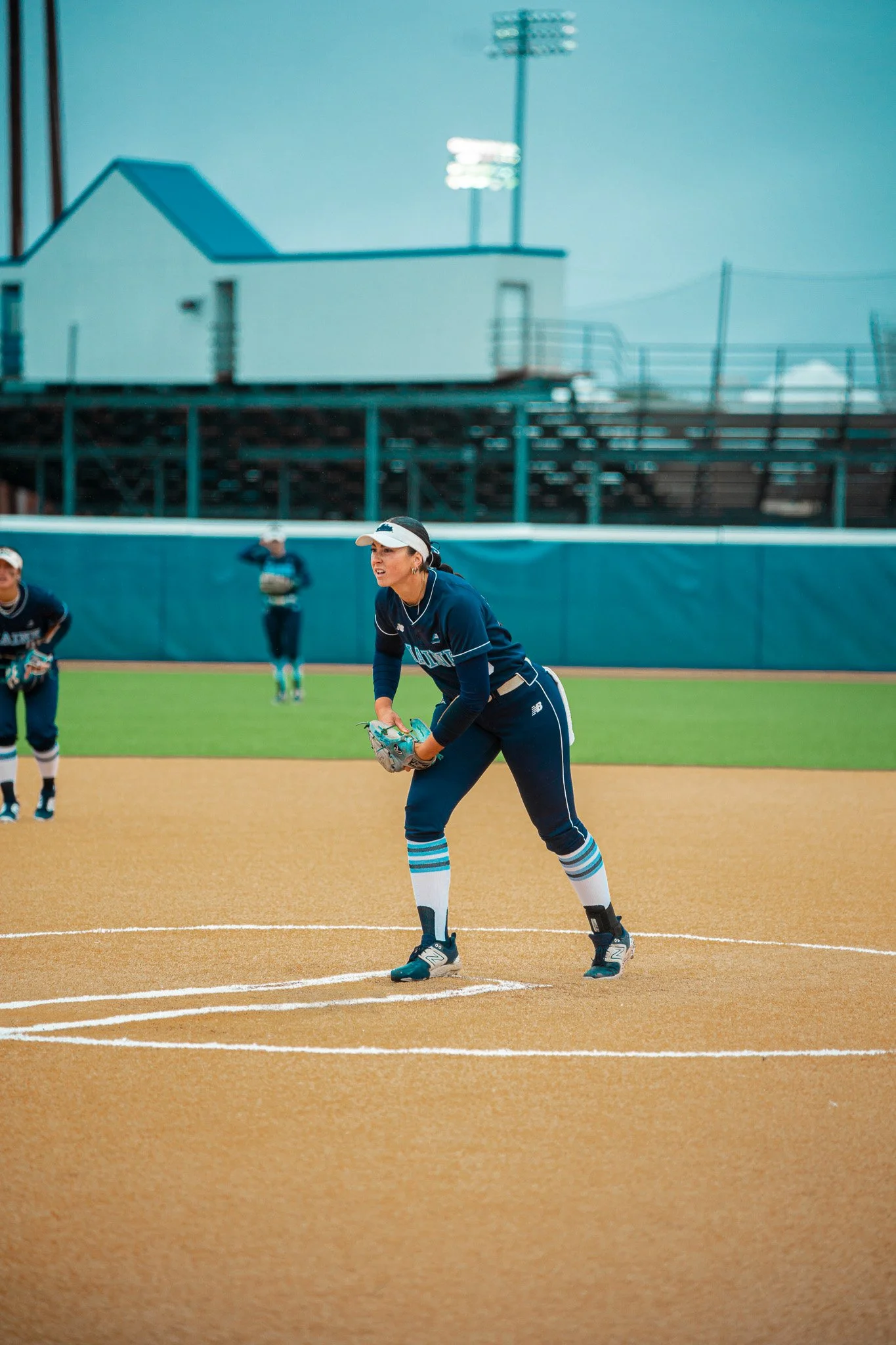 Softball player preparing to pitch on a field with teammates in the background, wearing a blue uniform.