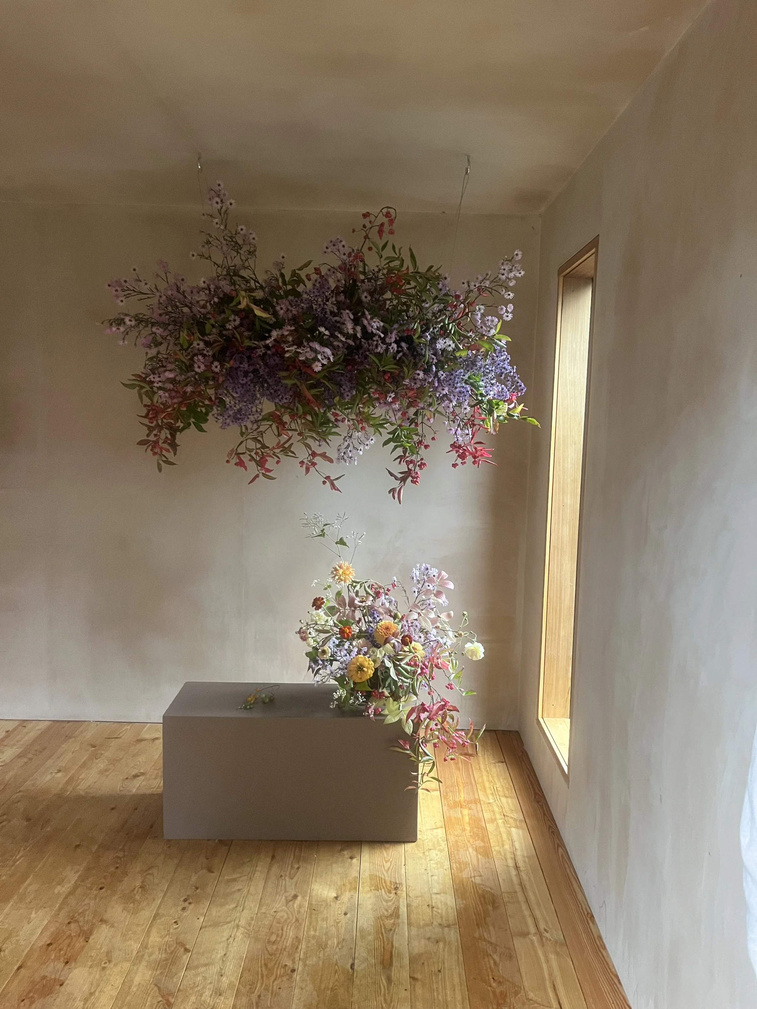 A floral arrangement with purple and pink flowers hangs from the ceiling, and a smaller floral arrangement with mixed flowers rests on a gray rectangular pedestal in a room with wooden flooring and a narrow window.