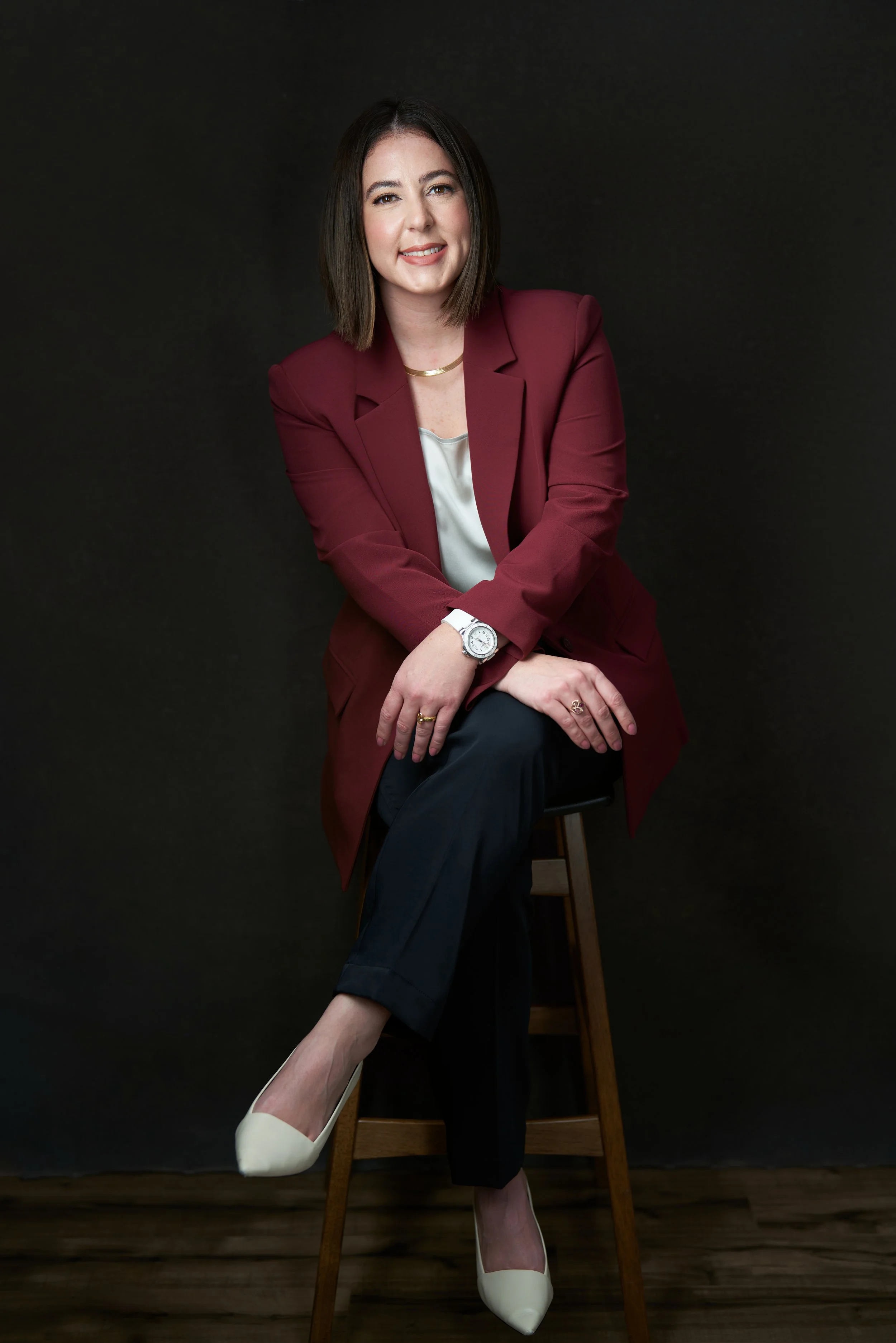 A woman with shoulder-length dark hair in a maroon blazer, cream blouse, black pants, and white high heels sitting on a wooden stool against a dark background.